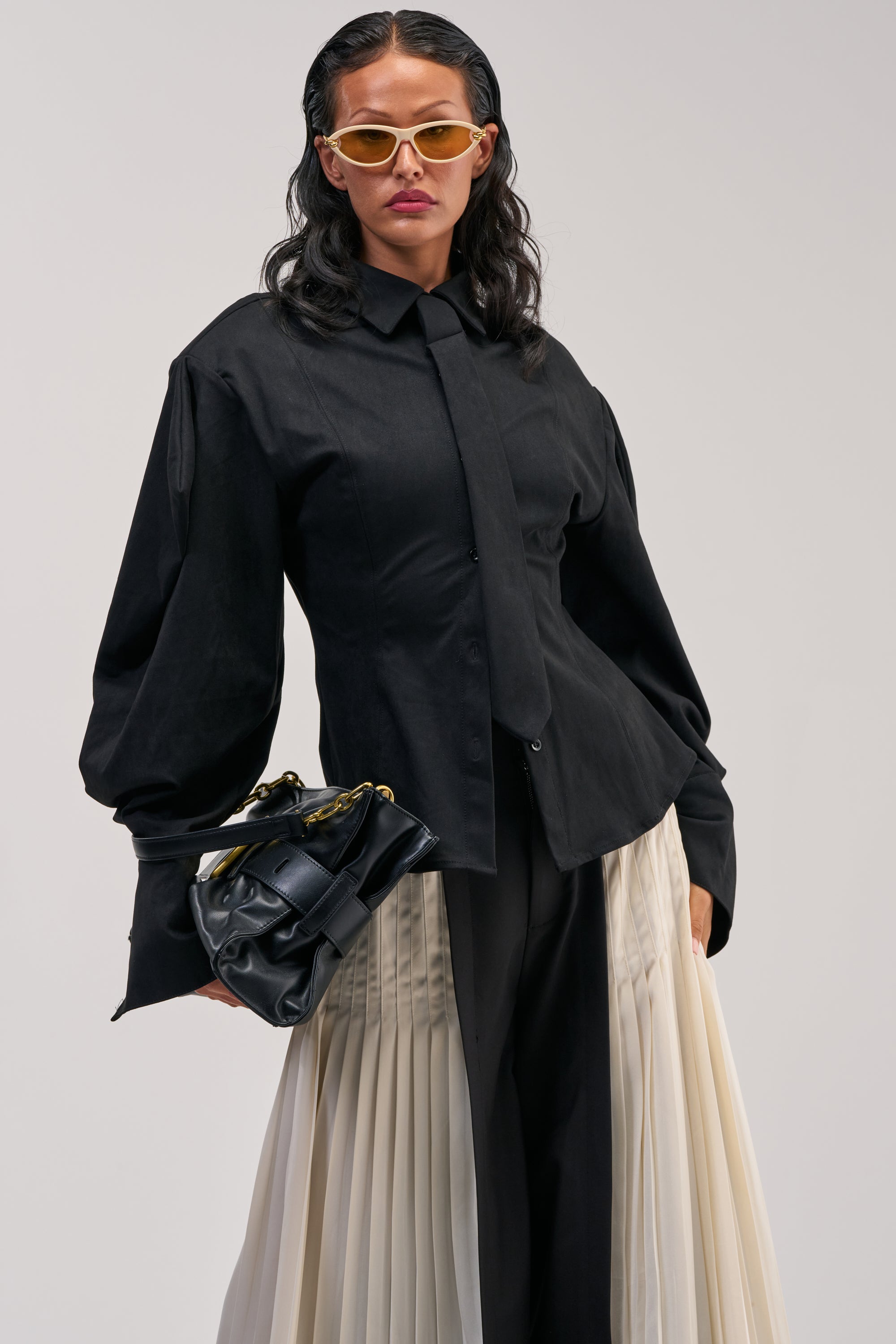 A person wearing the SHE MEANS BUSINESS SUEDE BUTTON DOWN with a matching tie, pleated cream and black skirt, yellow sunglasses, and holding a black handbag stands against a plain background.