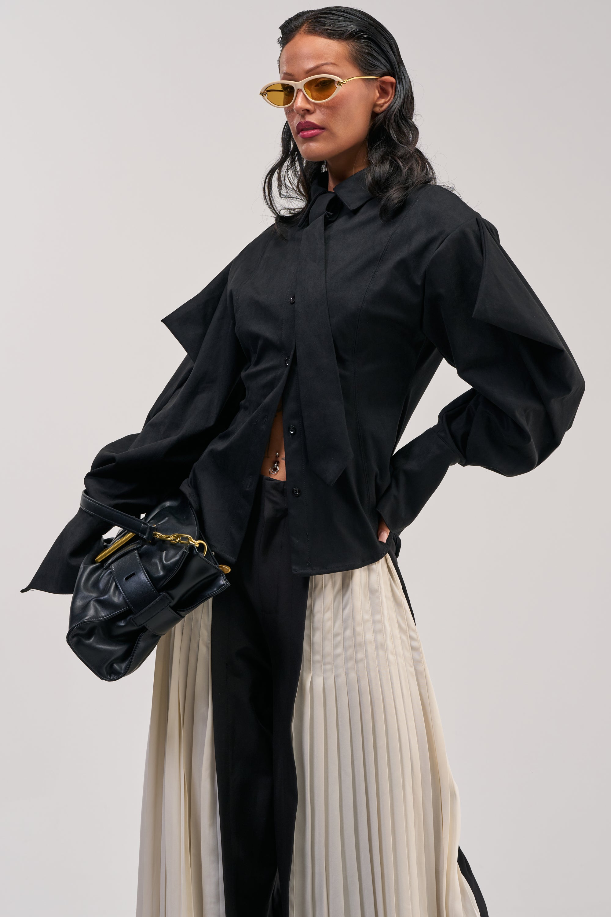 Wearing the SHE MEANS BUSINESS SUEDE BUTTON DOWN in black with oversized sleeves, a cream pleated skirt layered over black pants, yellow sunglasses, and carrying a black handbag, a person stands against a plain background.