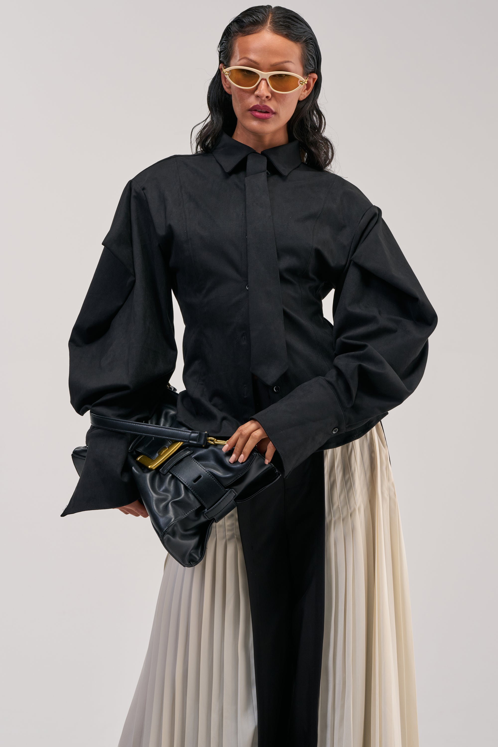 Wearing the SHE MEANS BUSINESS SUEDE BUTTON DOWN paired with a black tie, two-toned pleated skirt, yellow sunglasses, and carrying a black clutch bag, this individual poses confidently against a plain background.