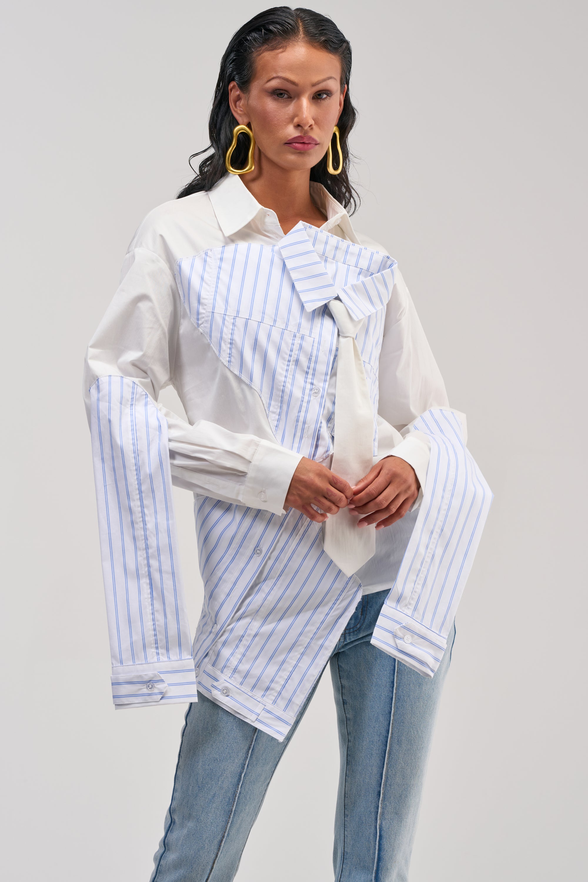 A woman wearing the HOT MESS POPLIN BLOUSE—a deconstructed white button-up with blue stripes and extra sleeves—paired with light blue jeans, yellow statement earrings, and slicked-back hair stands against a plain background.