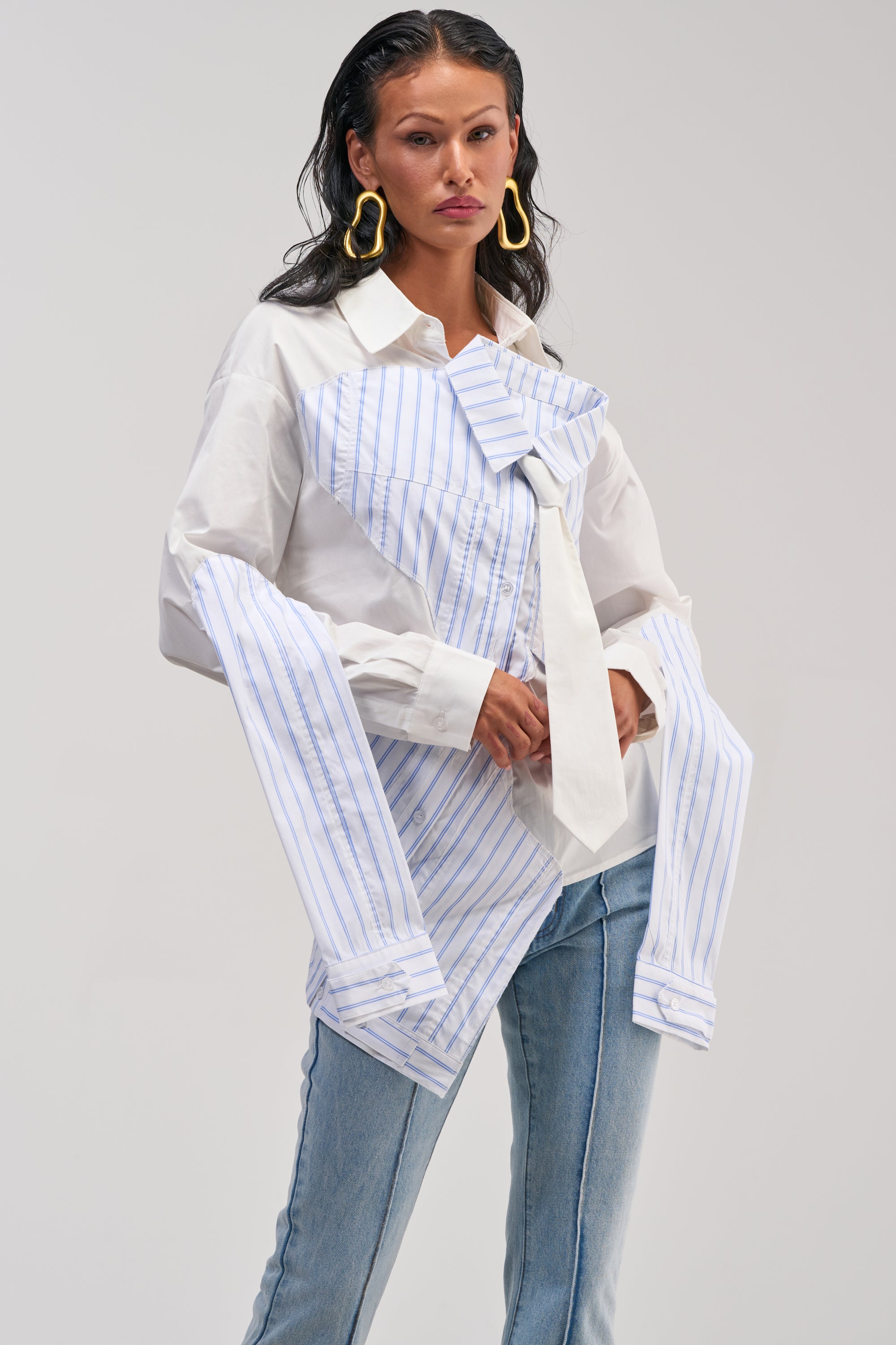A woman wears the HOT MESS POPLIN BLOUSE, a white button-up featuring an oversized blue-striped mock shirt and tie layered design, paired with light blue jeans and large gold earrings. She stands against a plain gray background.