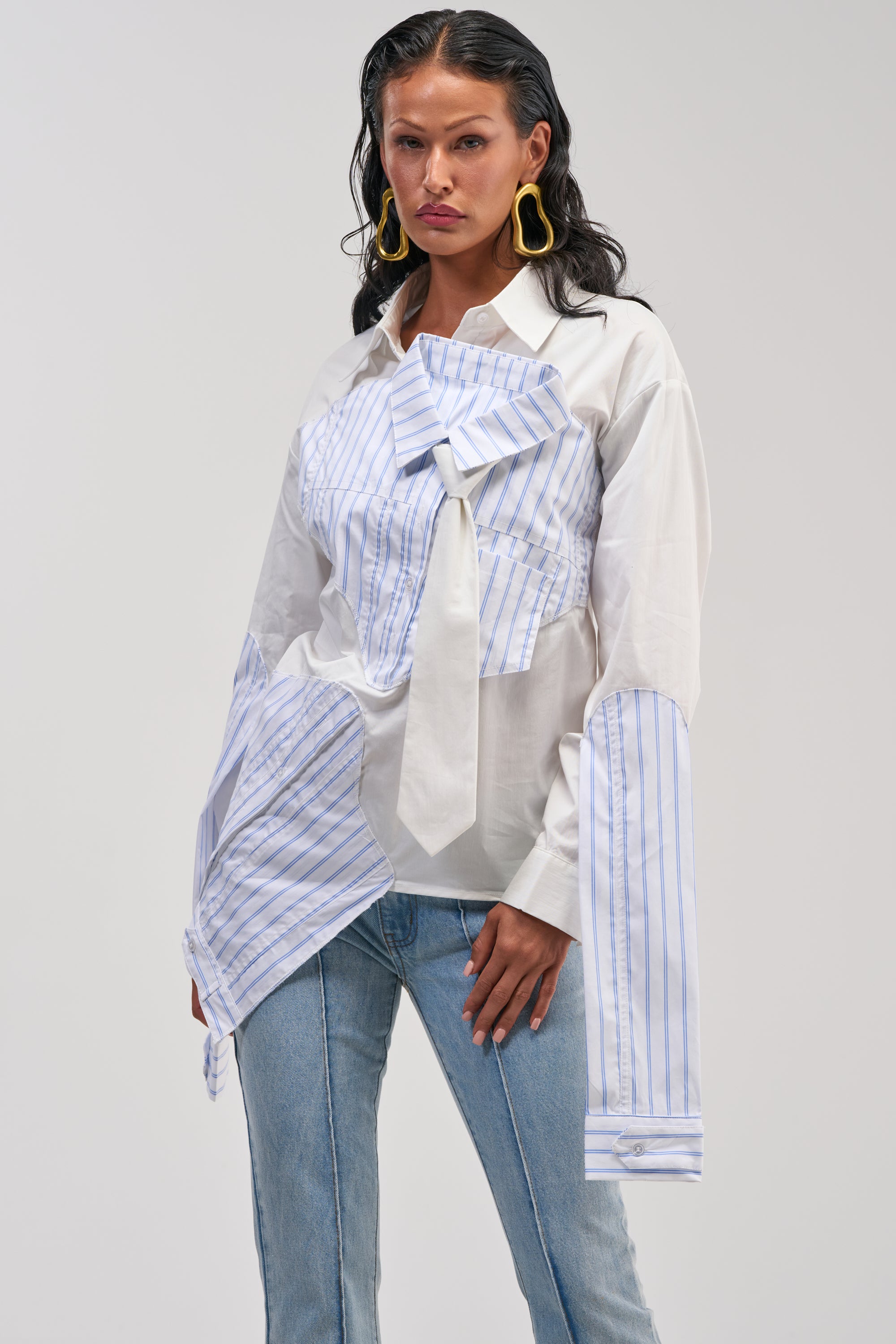 A woman poses against a plain background wearing the HOT MESS POPLIN BLOUSE, a deconstructed white button-up featuring asymmetrical blue-striped panels, oversized cuffs, and a bow detail. She pairs it with light blue jeans, accessorizing with large yellow statement earrings and sporting wet-look hair.