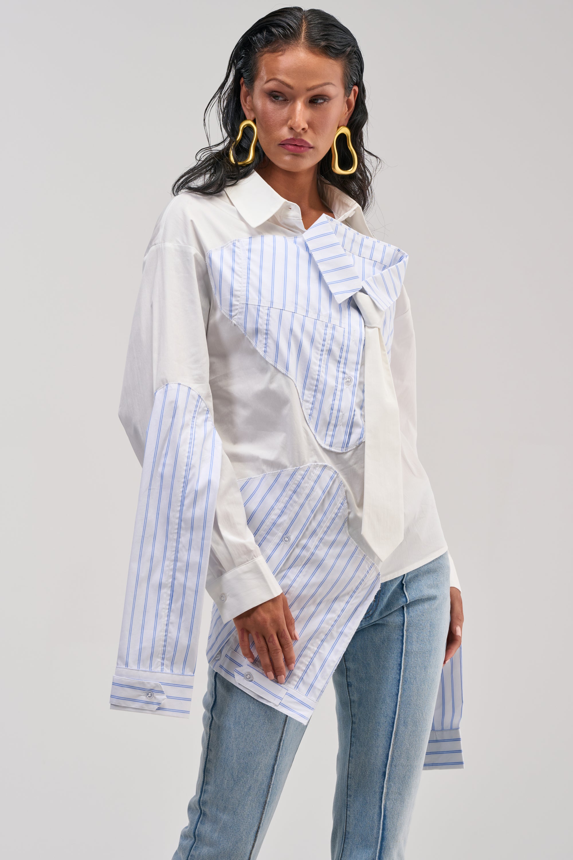 A woman wears the HOT MESS POPLIN BLOUSE, a deconstructed white button-up with blue striped patches, oversized cuffs, and a corseted back panel. She pairs it with light blue jeans and large gold earrings, standing against a plain gray background.