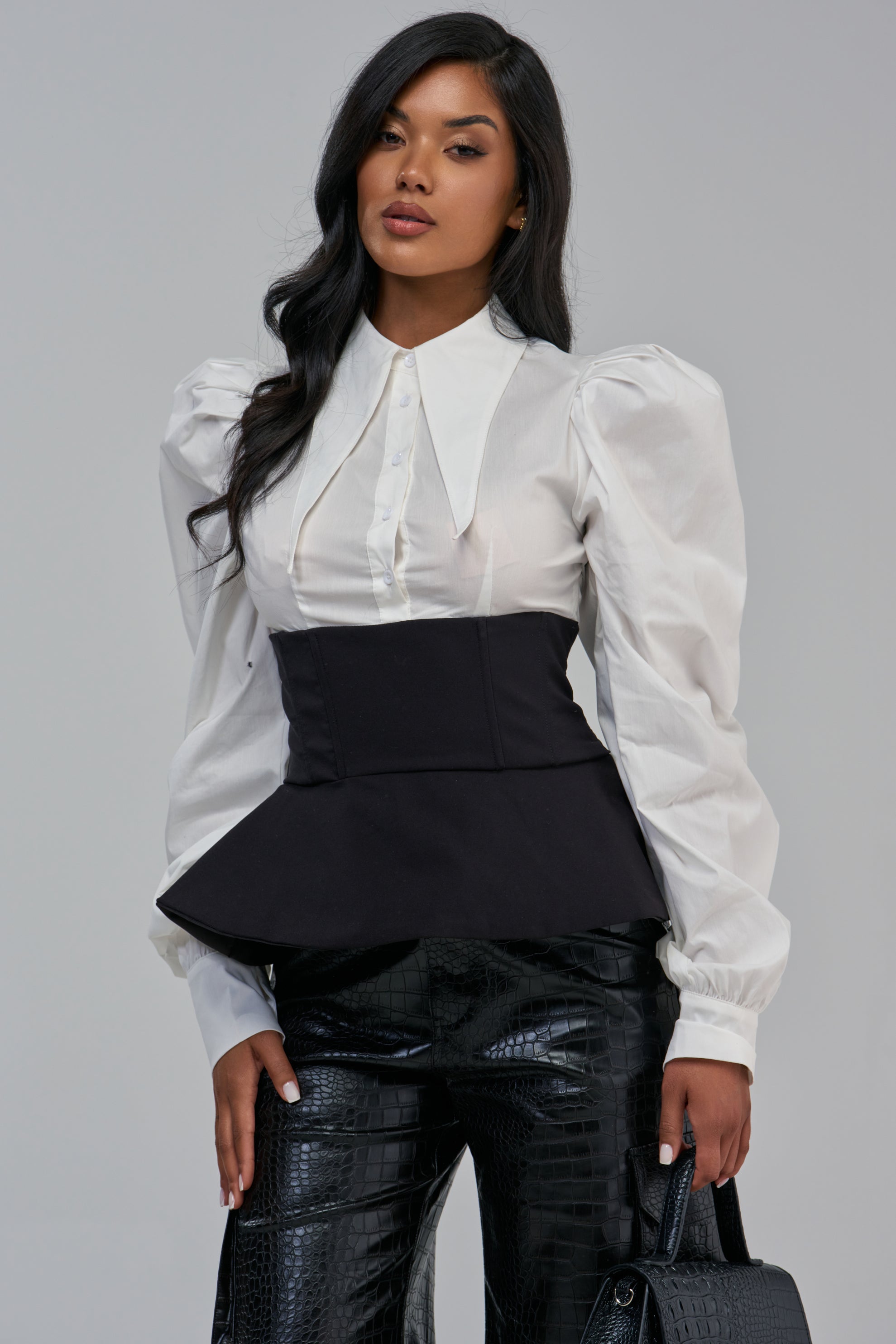 A person wearing the JULIETTE CORSETTED BLOUSE, a white long-sleeved peplum blouse with a pointed collar and puff shoulders, layered under a black corset-style waist.