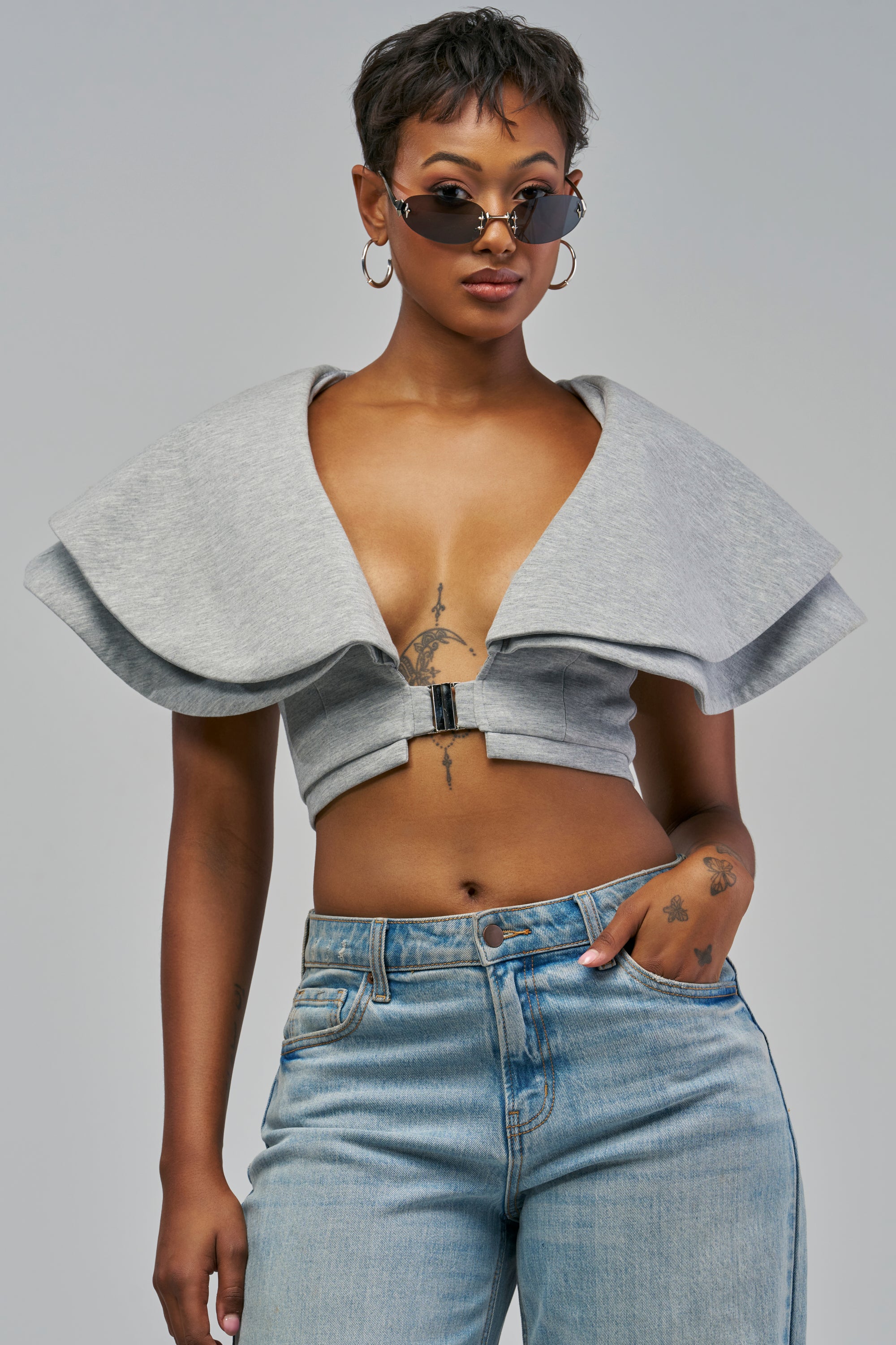 The MAXIMALIST SCUBA TOP is a grey scuba batwing crop top with a deep V-neck and wide, layered lapels, worn with blue jeans for an avant-garde look.