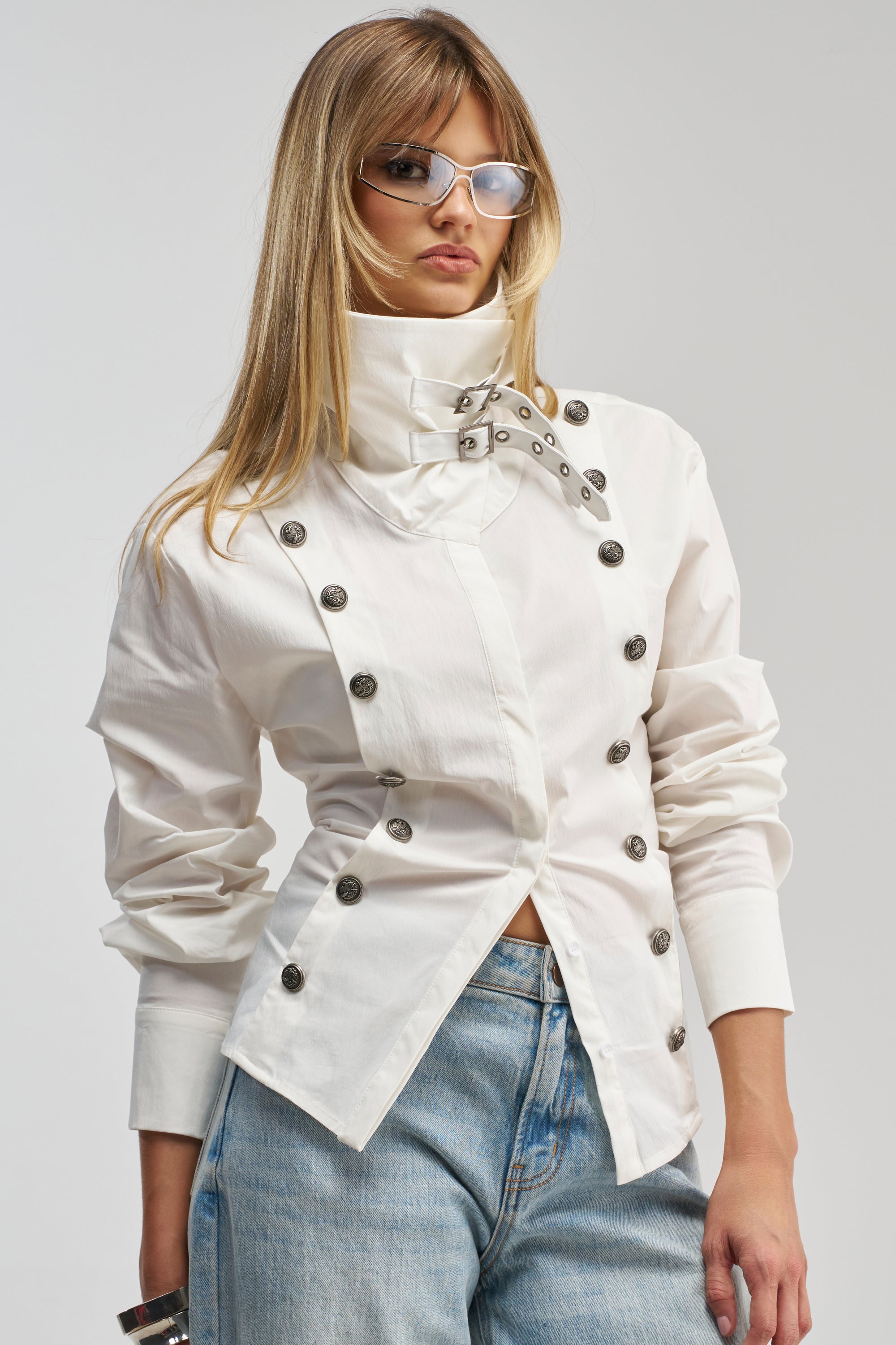 A woman in blue jeans and the IN YOUR DREAMS BUTTON DOWN—a crisp white shirt featuring metallic buttons and a high collar—wears sunglasses and stands against a plain background for an effortlessly chic take on modern officewear.