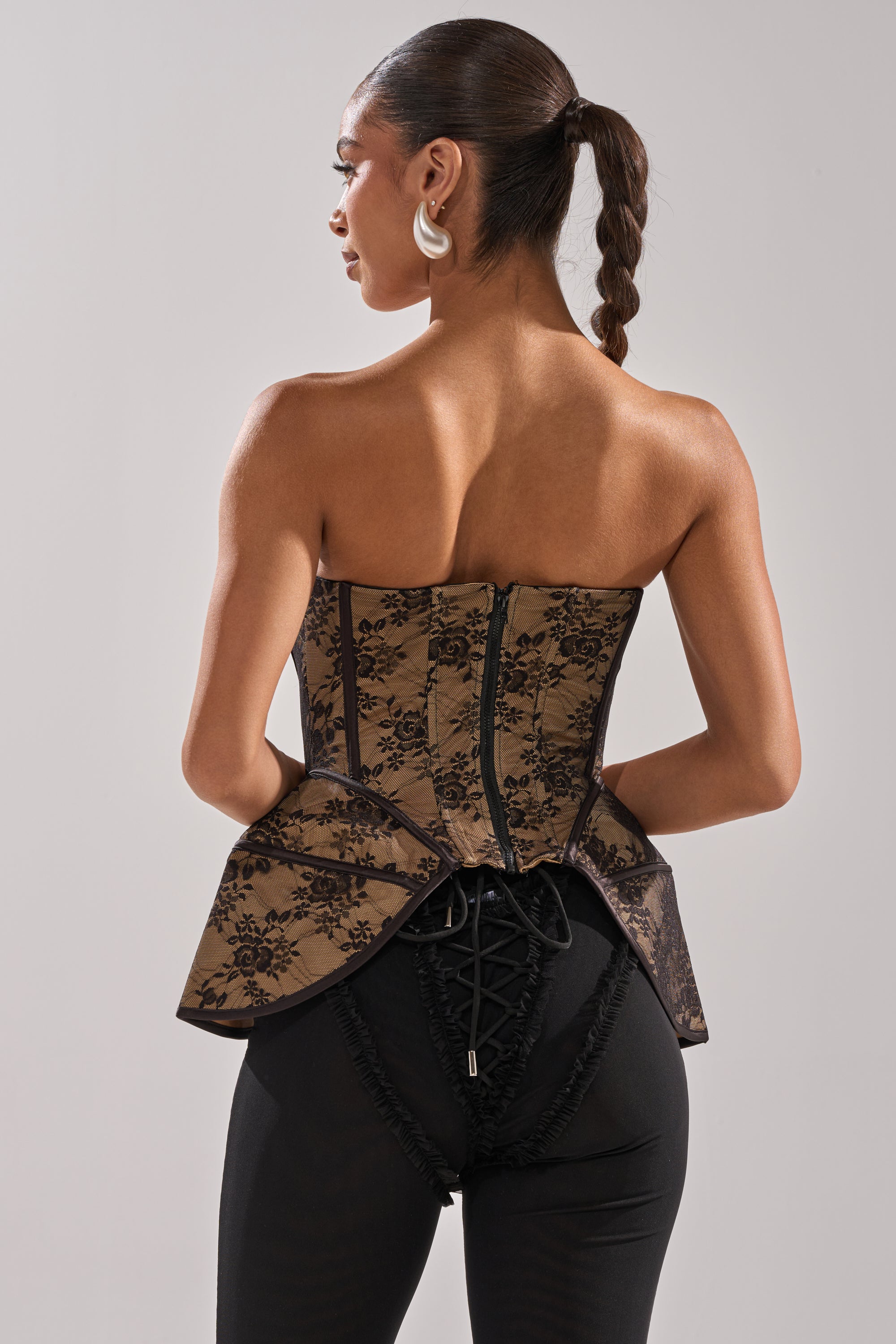 A woman with a braided ponytail, facing away from the camera, showcases the lace-up and zipper details on the back of her elegant JUST A GIRL LACE CORSET paired with black pants.