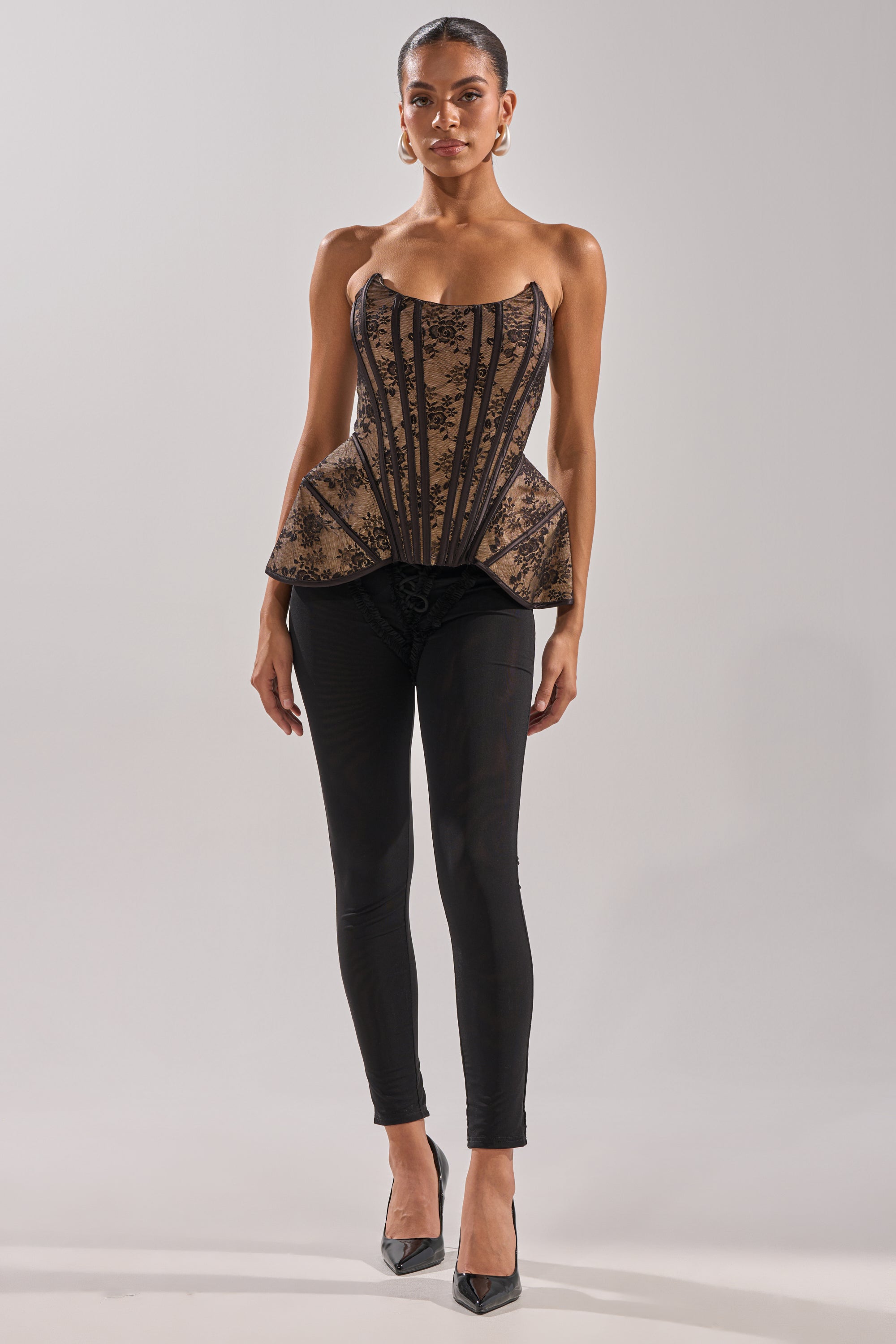 A woman wears the JUST A GIRL LACE CORSET, a strapless black floral piece, paired with black pants, hoop earrings, pointed black heels, and styled hair pulled back, all set against a plain light background.