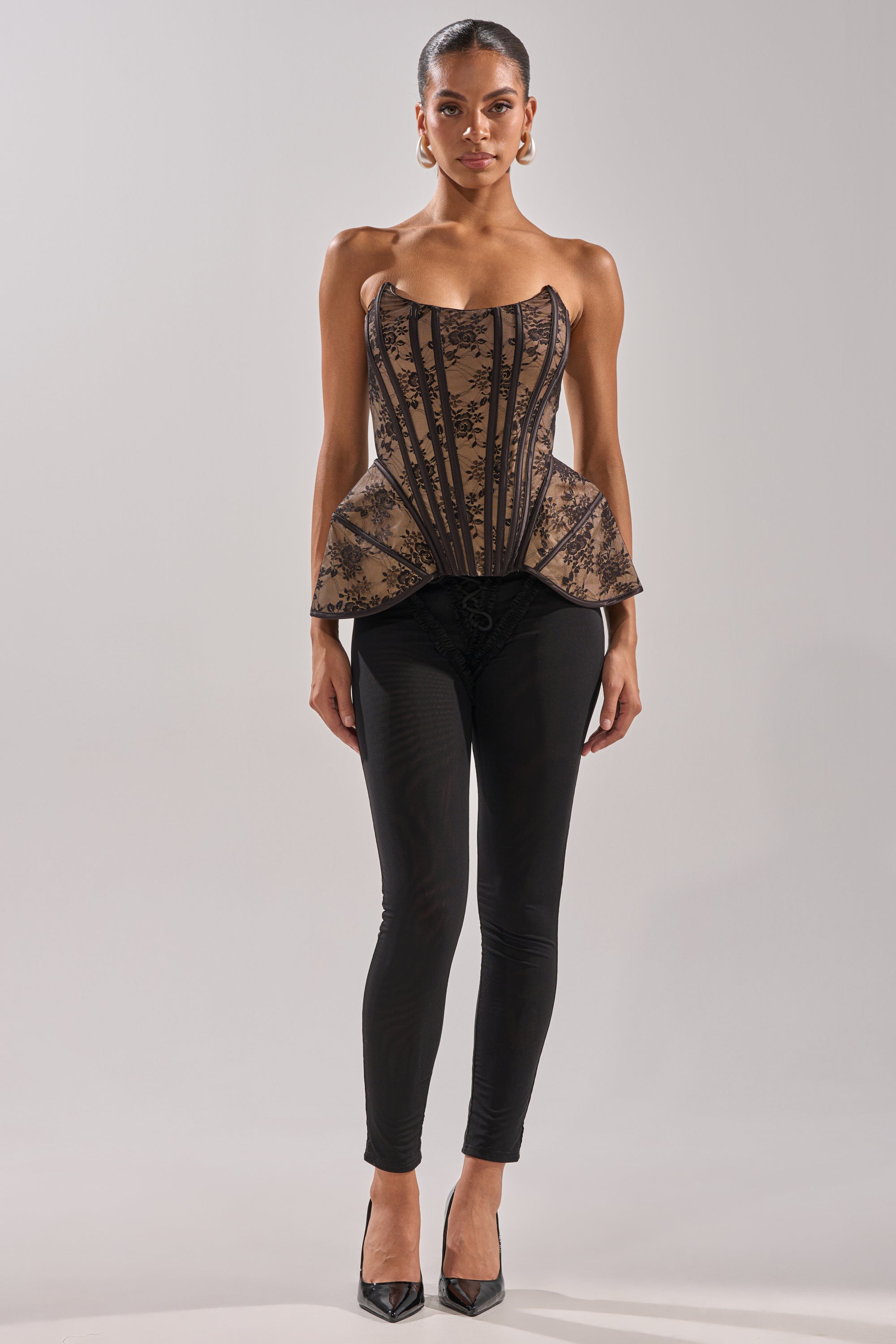 A woman faces forward in the JUST A GIRL LACE CORSET, a strapless black floral corset with a structured beige base and black lace overlay, styled with black fitted pants and high heels against a plain background.