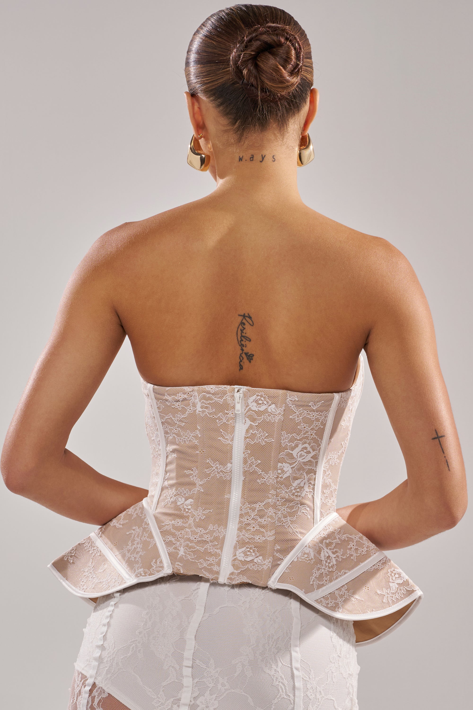 A woman with tattoos on her upper back, neck, and left arm stands against a light background wearing the JUST A GIRL LACE CORSET IN WHITE; her hair is styled in a neat bun and she is seen from behind.