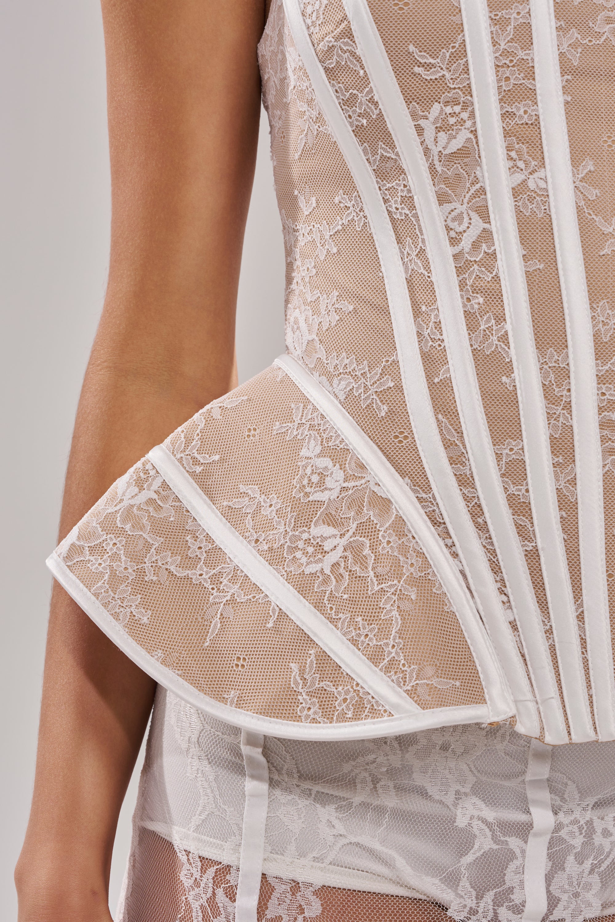 A close-up of a person wearing the JUST A GIRL LACE CORSET IN WHITE, featuring sheer fabric, boning, a peplum hem, delicate floral patterns, intricate stitching, and matching lace underwear.