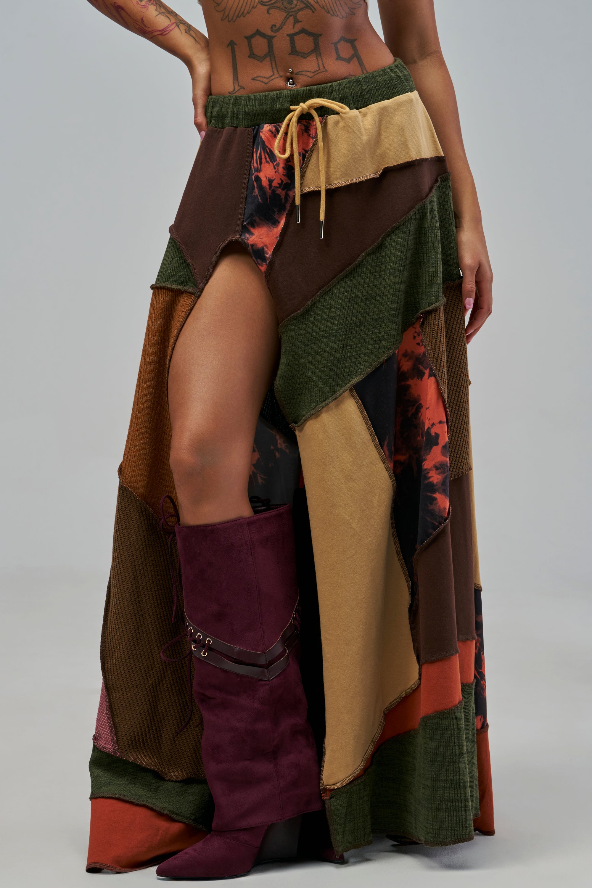 A person wears an ALARA PATCHWORK MAXI SKIRT with a drawstring waist and a high front slit, styled in boho streetwear fashion with a deep red knee-high boot.