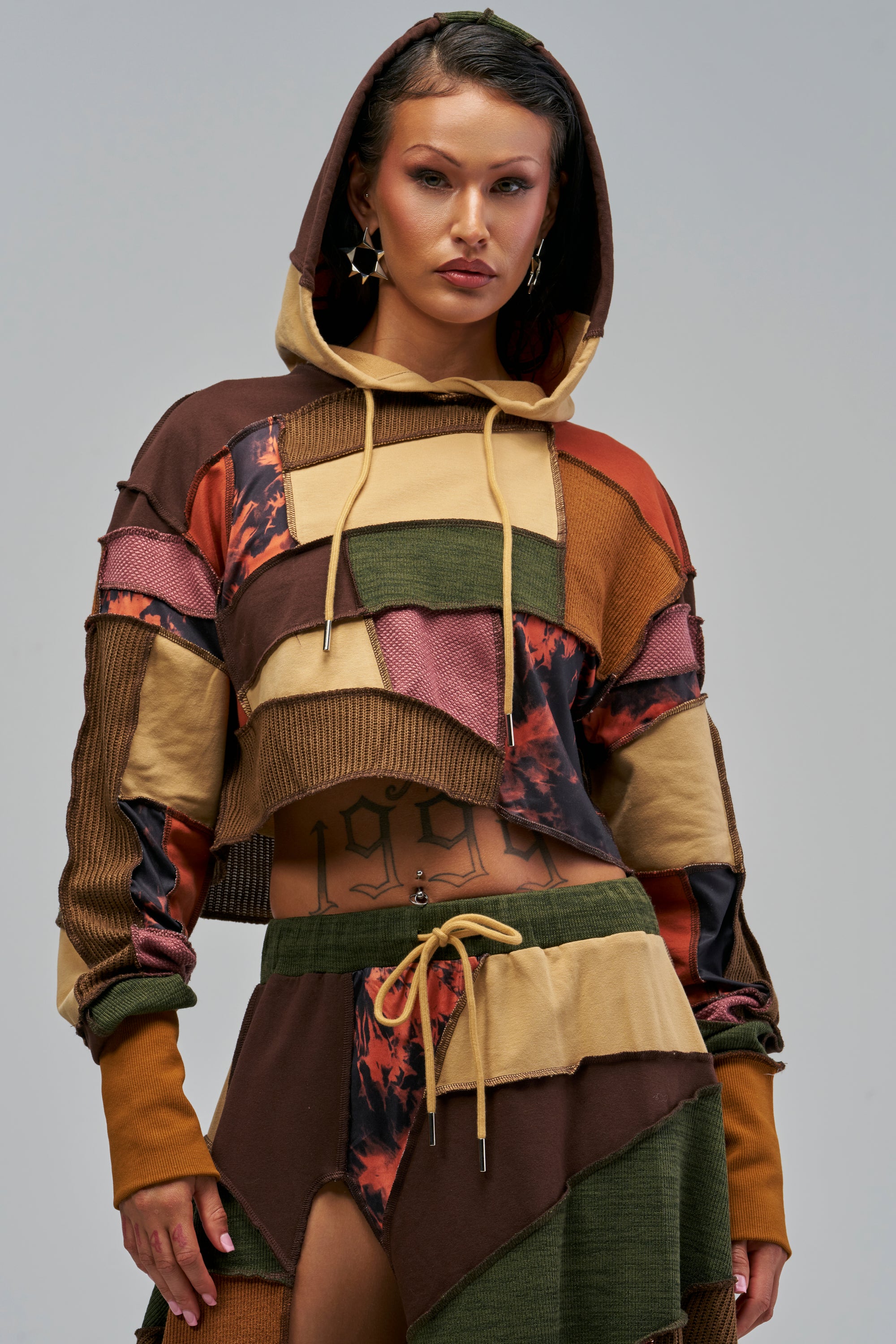 A person wears an ALARA PATCHWORK SWEATSHIRT cropped hoodie made of various brown, tan, and green fabric patches, paired with matching patchwork joggers.