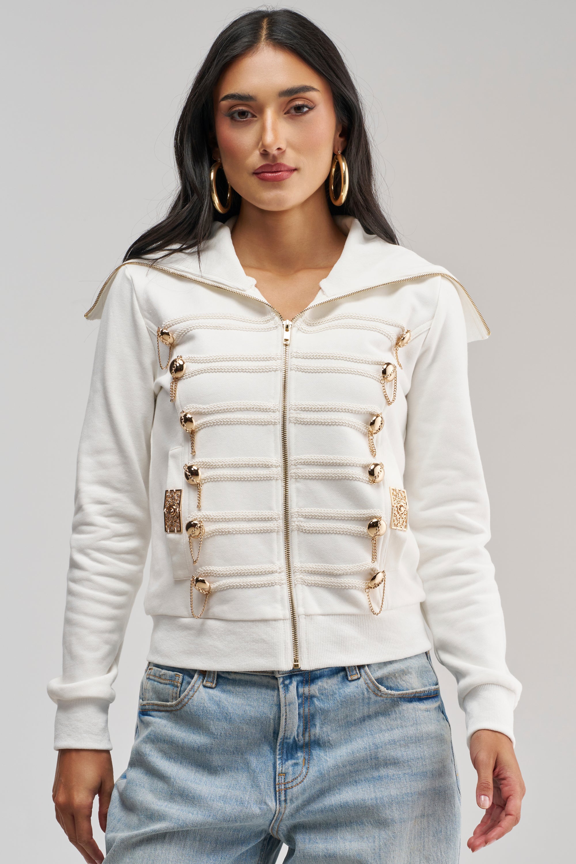 A woman stands facing forward against a plain background, layering the OUT OF YOUR LEAGUE EMBELLISHED SWEATSHIRT—with decorative gold safety pins—over blue jeans for an effortlessly utility-chic streetwear look.