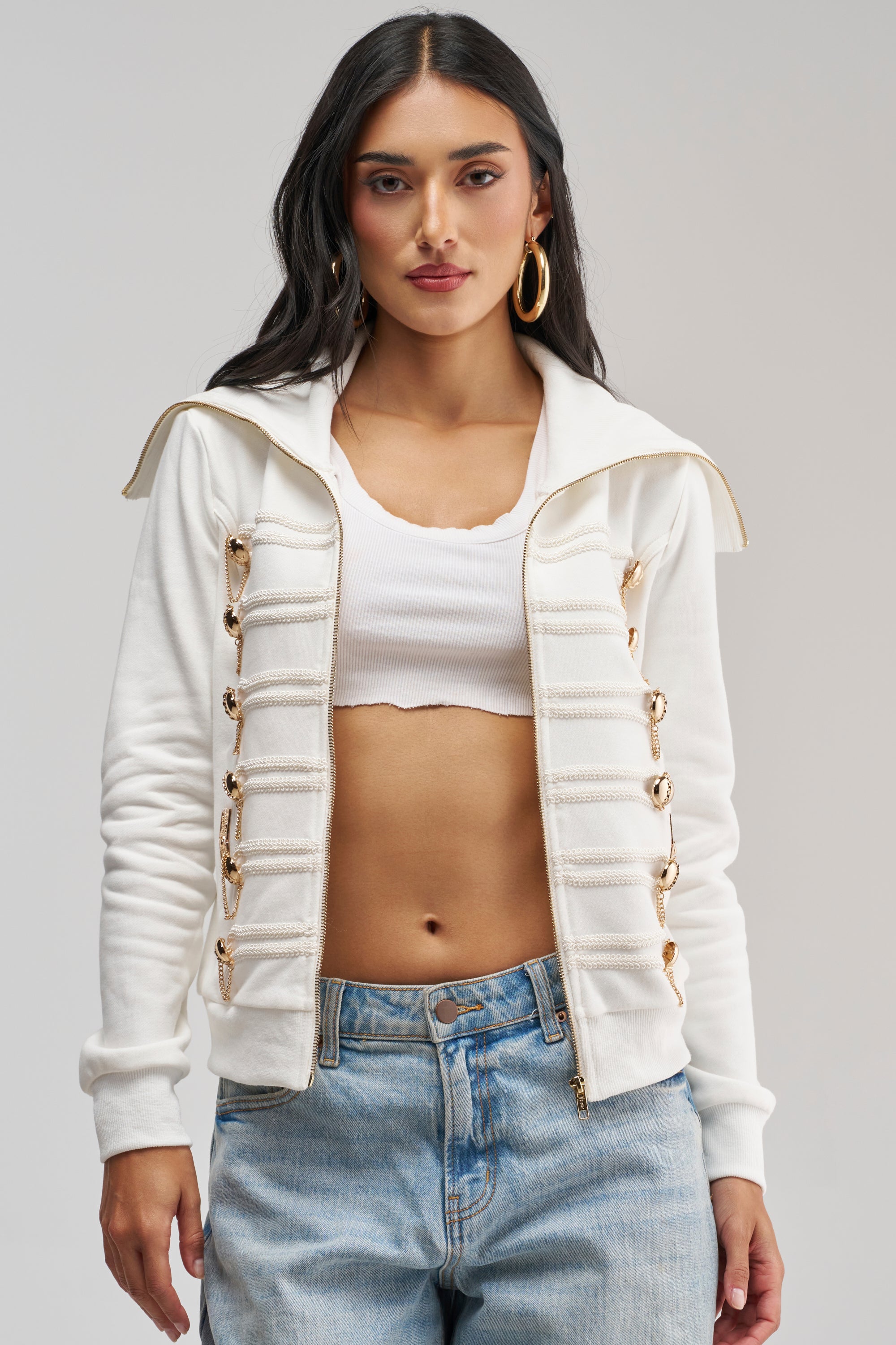 A woman stands against a light gray background, showcasing utility-chic style in the OUT OF YOUR LEAGUE EMBELLISHED SWEATSHIRT—a military-inspired zip-up piece with gold accents—layered over a white crop top and paired with light blue jeans.