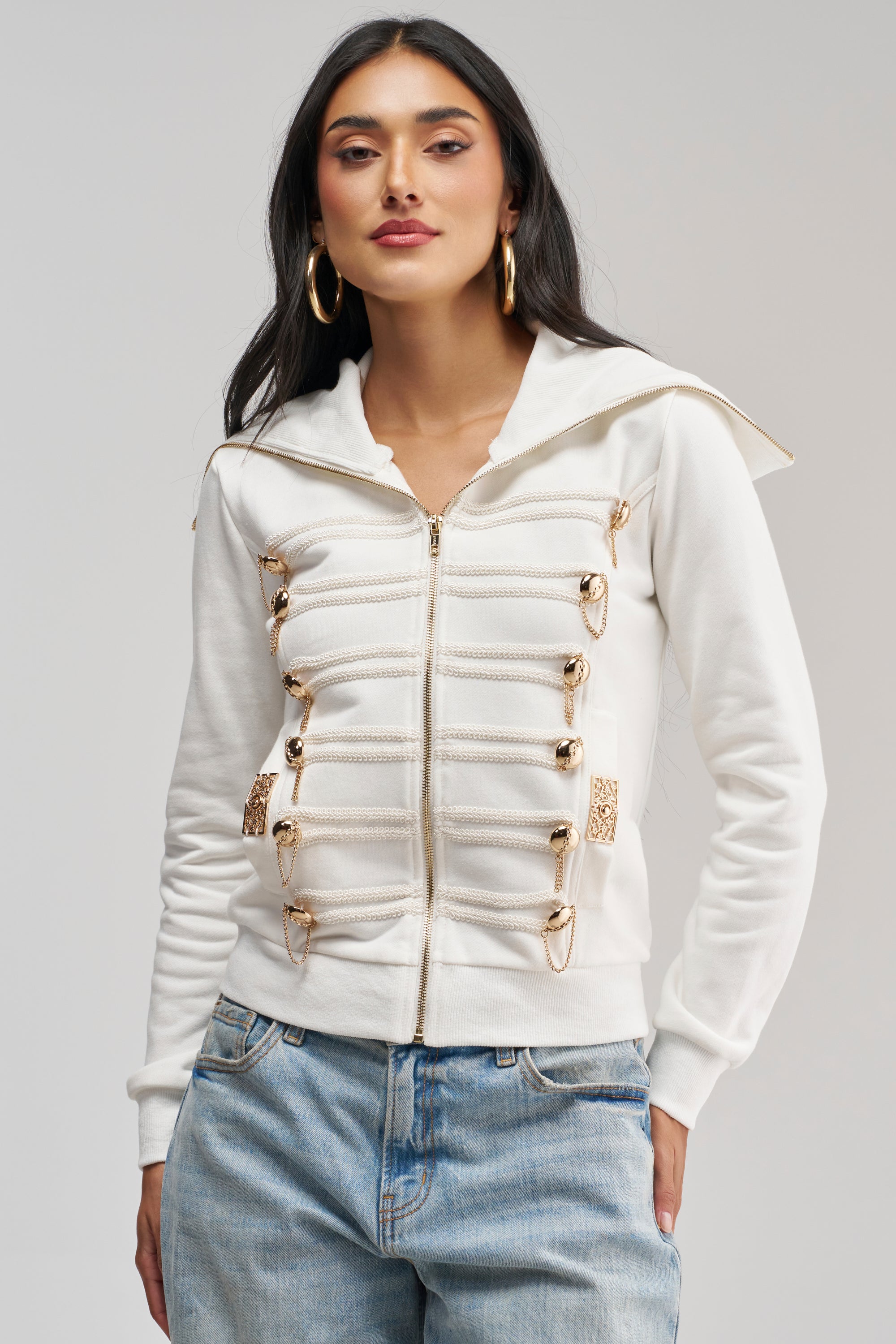 Wearing the OUT OF YOUR LEAGUE EMBELLISHED SWEATSHIRT—a white, military-inspired zip-up with gold decorative clasps—paired with blue jeans and set against a plain light background, this look is a flawless example of utility-chic style for easy streetwear layering.