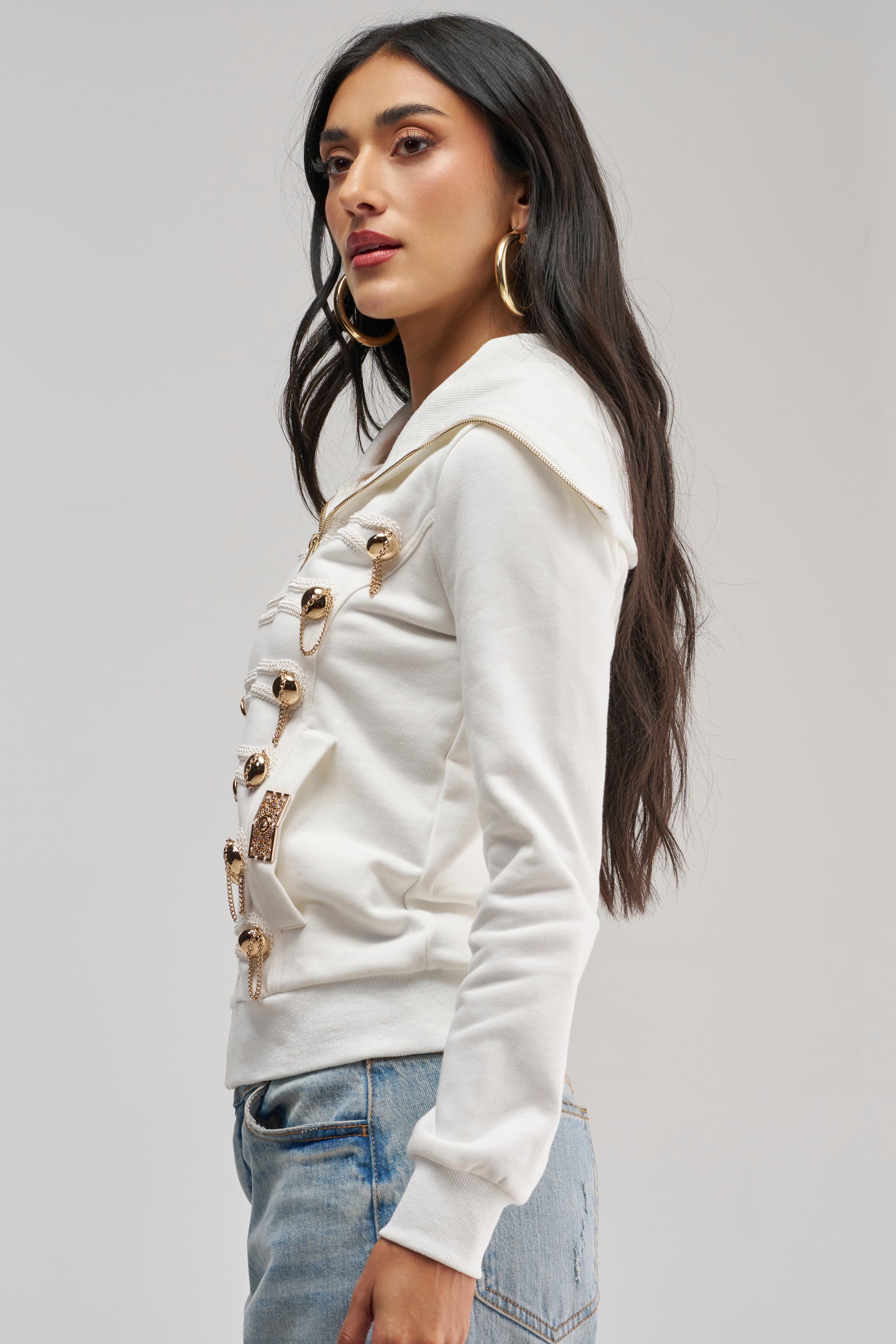 A woman with long dark hair stands in profile against a light gray background, effortlessly showcasing utility-chic style in the OUT OF YOUR LEAGUE EMBELLISHED SWEATSHIRT—a white, military-inspired zip-up design adorned with gold decorative buttons—paired with blue jeans.