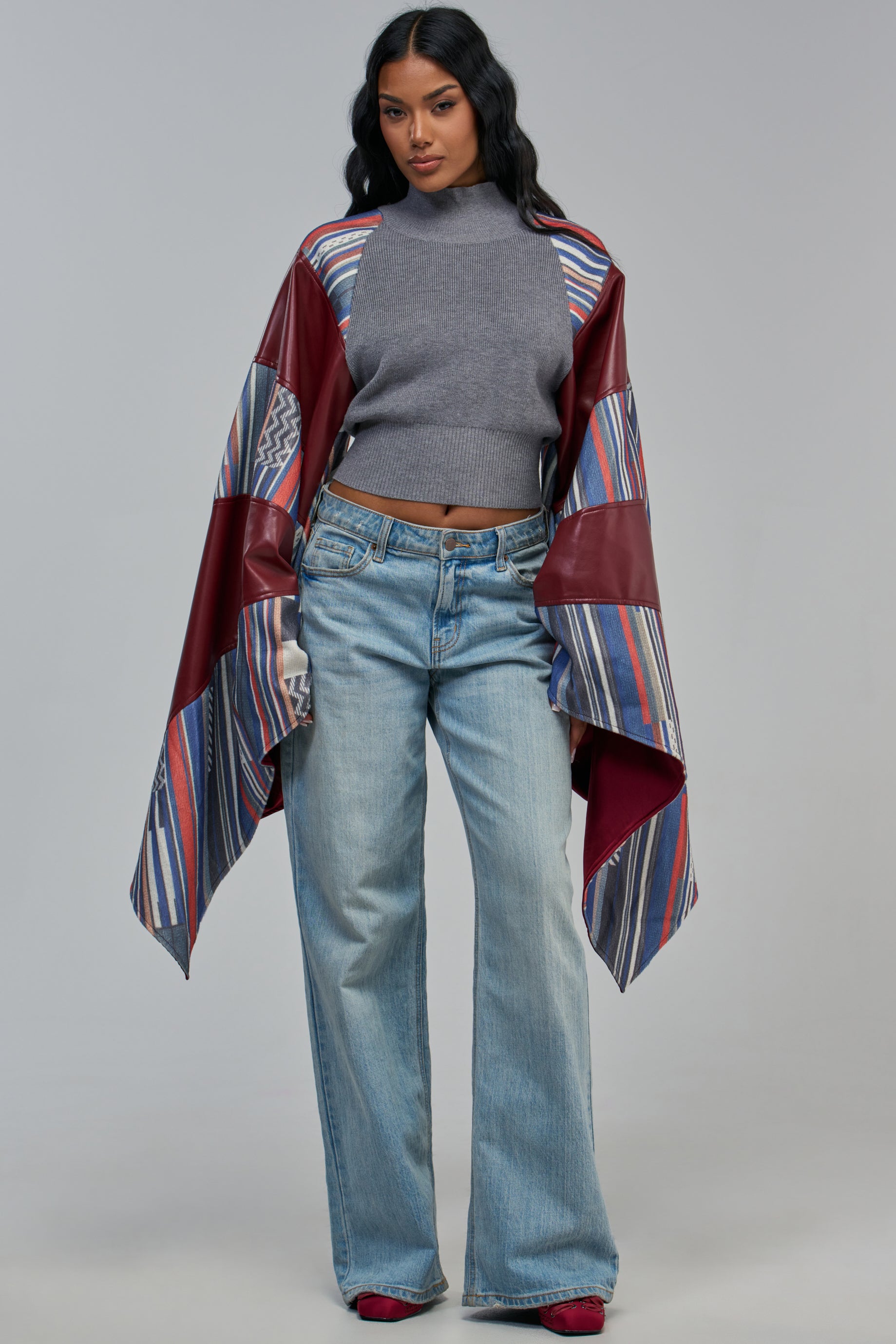 A woman wears a CATCHING A VIBE CROPPED SWEATER with billowing sleeves under a draped, open-front garment with red faux leather and striped panels, paired with light-wash wide-leg jeans.