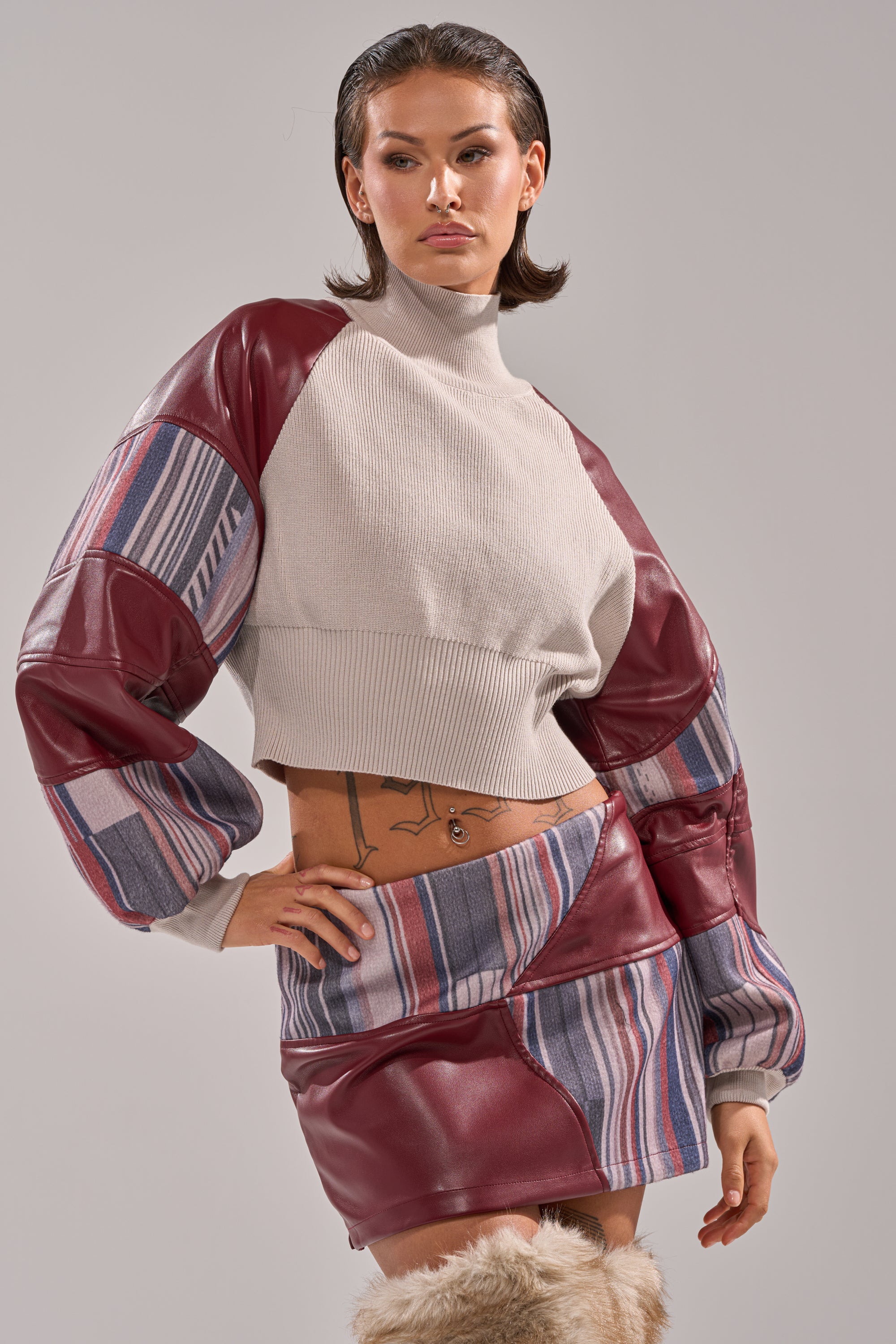 A person poses confidently in the FEELING THE VIBES CROPPED GREY SWEATER, matching mini skirt, and knee-high furry boots against a plain background. 0
