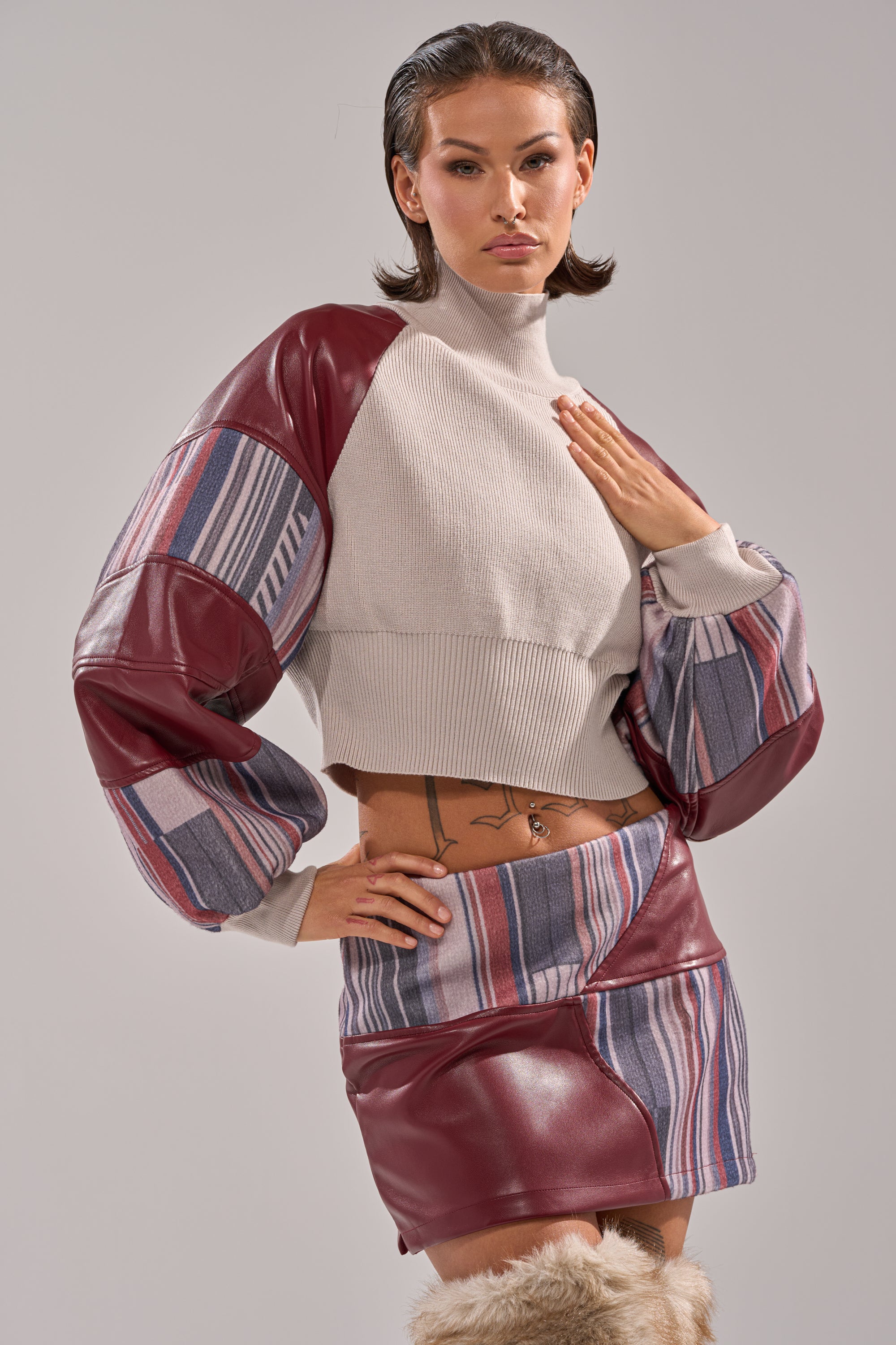 A woman poses confidently in the FEELING THE VIBES CROPPED GREY SWEATER with voluminous sleeves, a matching mini skirt with maroon and striped panels, and knee-high fur boots against a neutral background. 1