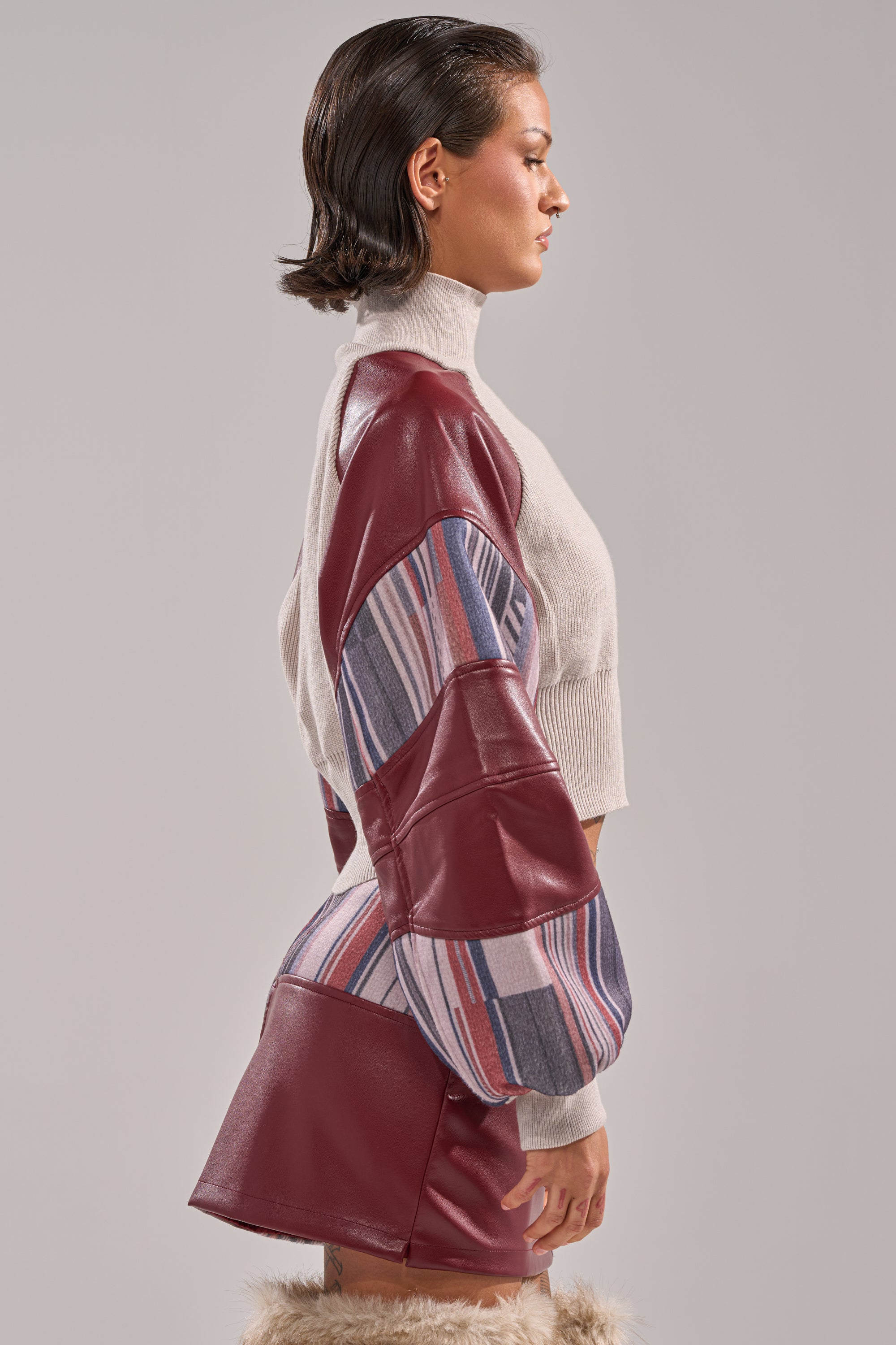 A woman stands in profile against a plain gray background, wearing the FEELING THE VIBES CROPPED GREY SWEATER paired with a maroon faux leather and multicolored striped skirt and fur-trimmed boots. 2