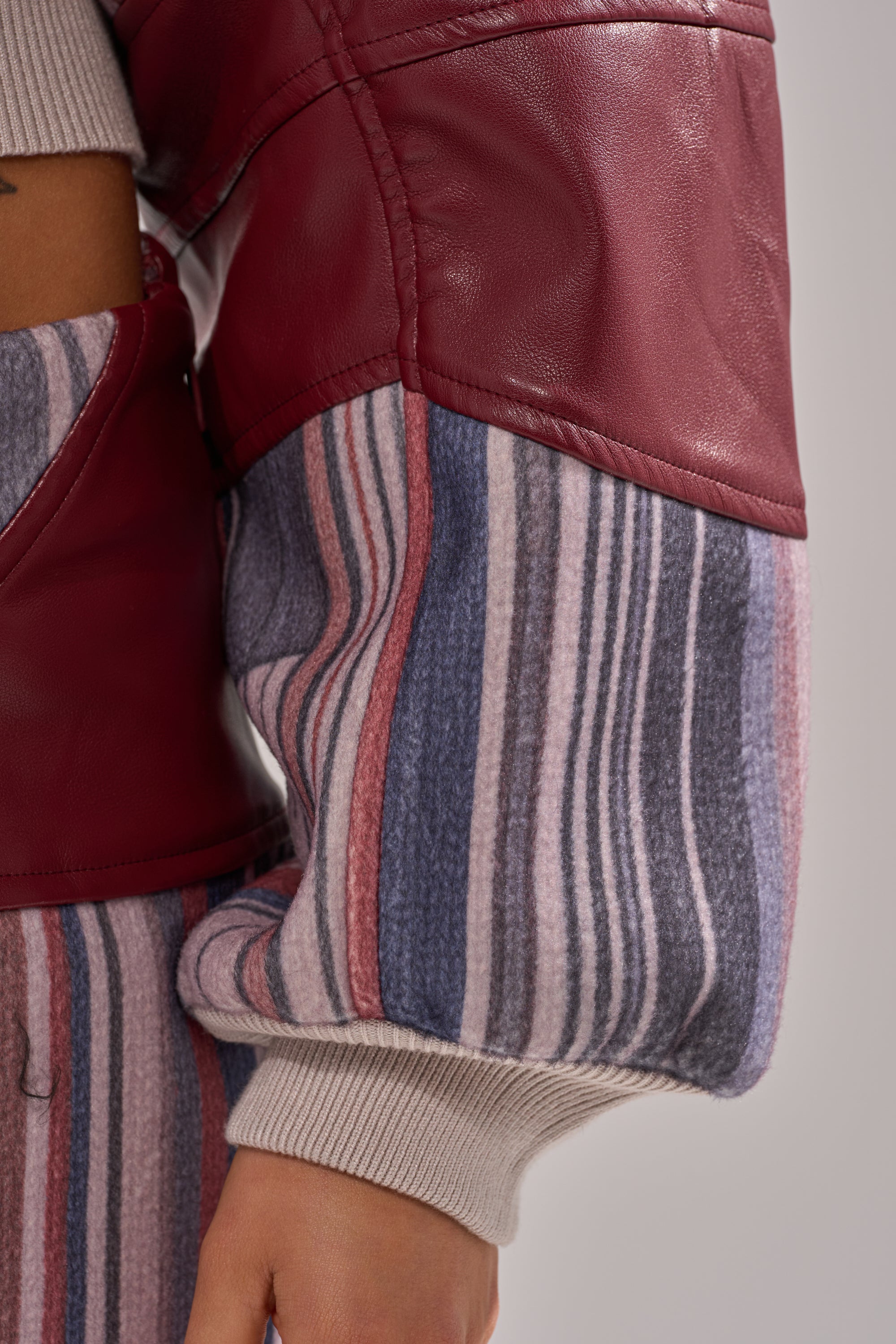 A close-up of a person's arm wearing the FEELING THE VIBES CROPPED GREY SWEATER, featuring colorblock sleeves with a maroon leather upper and soft, striped fabric in gray, pink, blue, and white. The hand is partially visible. 7