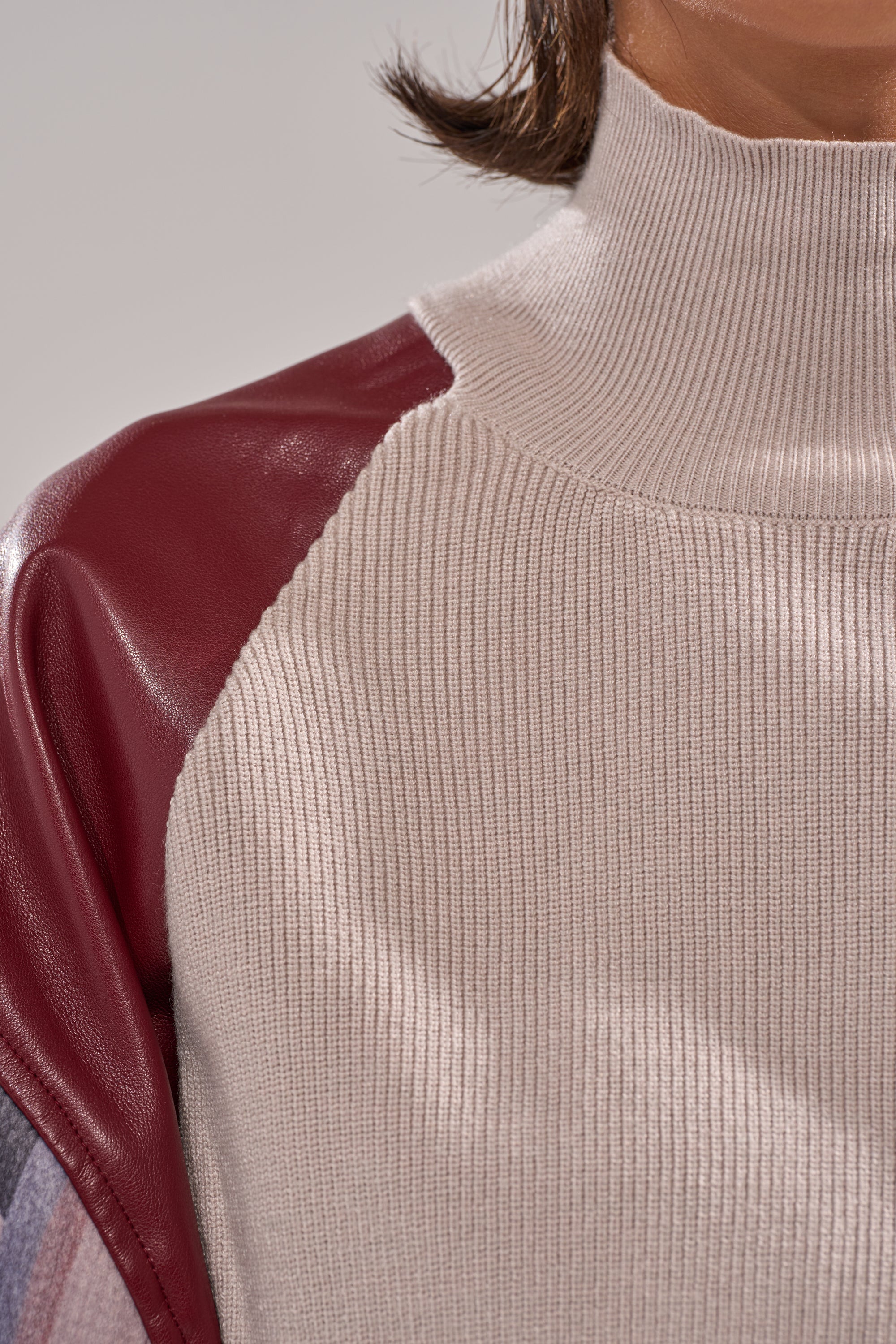 Close-up of a person wearing the FEELING THE VIBES CROPPED GREY SWEATER, featuring a light beige ribbed knit body and contrasting maroon faux leather colorblock sleeves. The image highlights fabric texture and contrasting materials. 8