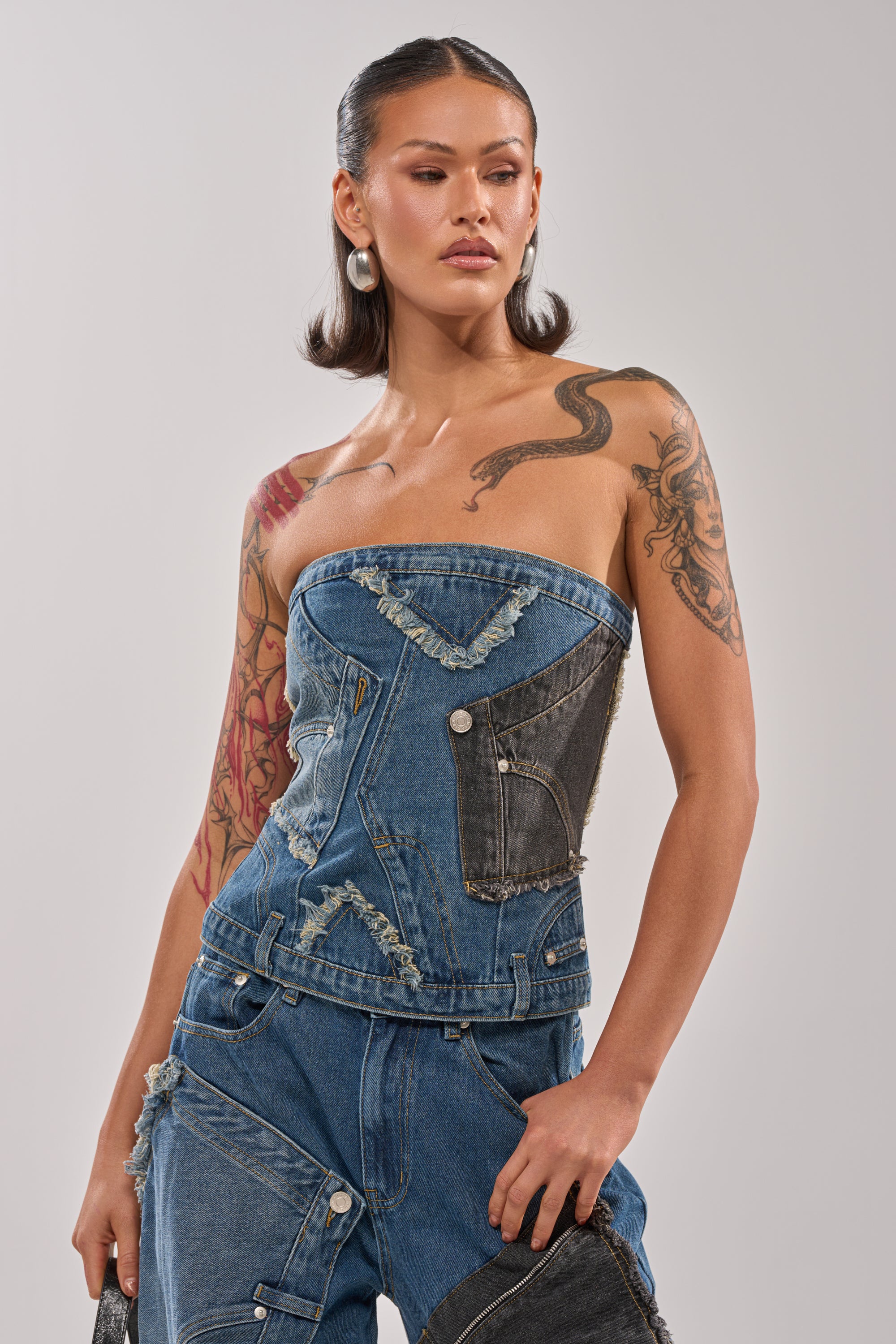 A woman with a sleek bob models the MIXED MATCH DENIM CORSET top with matching jeans, silver hoop earrings, and visible tattoos. She poses against a plain light background, channeling bold street style. 0