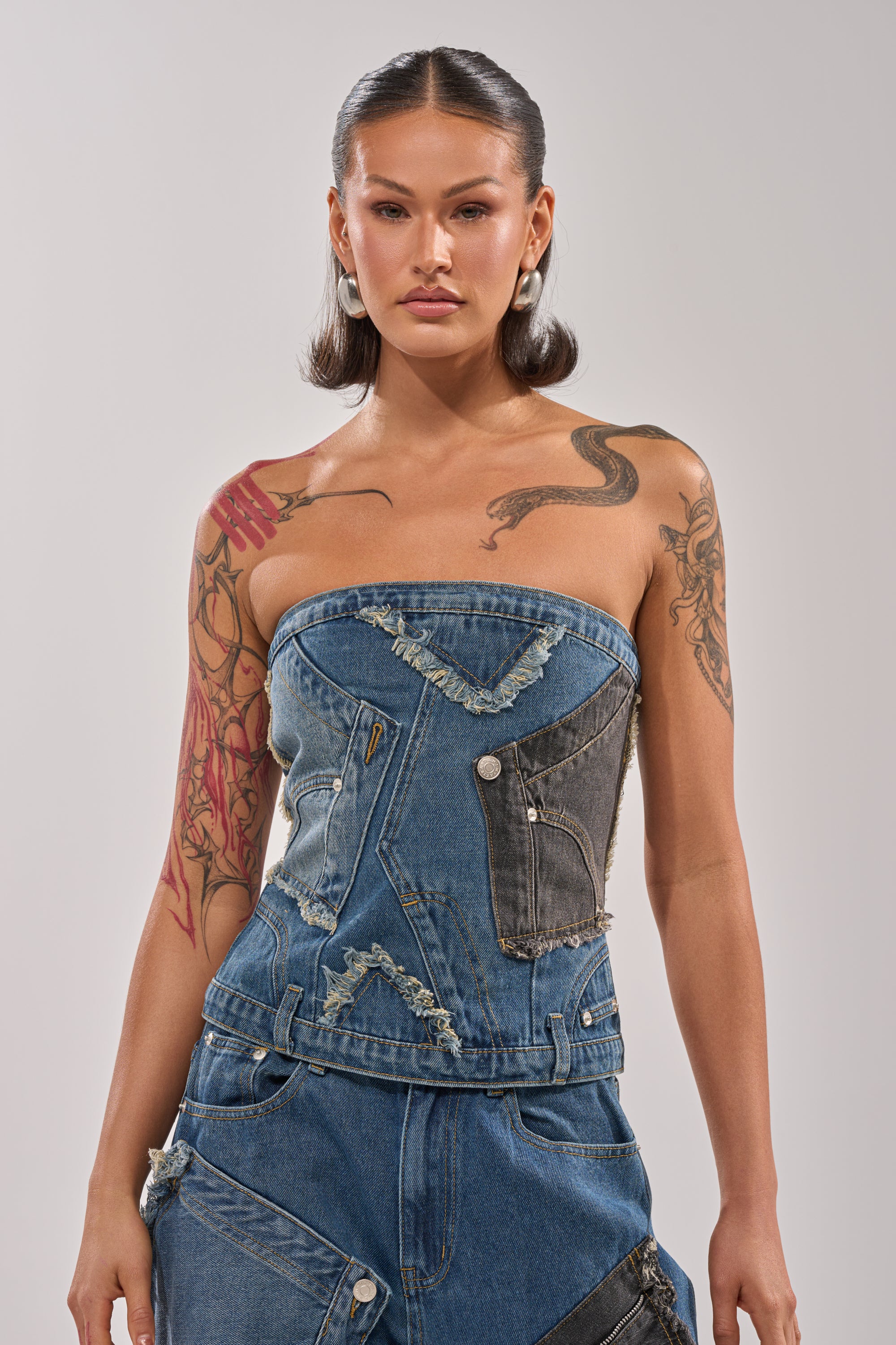A woman with tattoos rocks street style in the MIXED MATCH DENIM CORSET and patchwork, frayed-edge denim pants. Her sleek, medium-length dark hair and large earrings stand out against a plain light background. 1