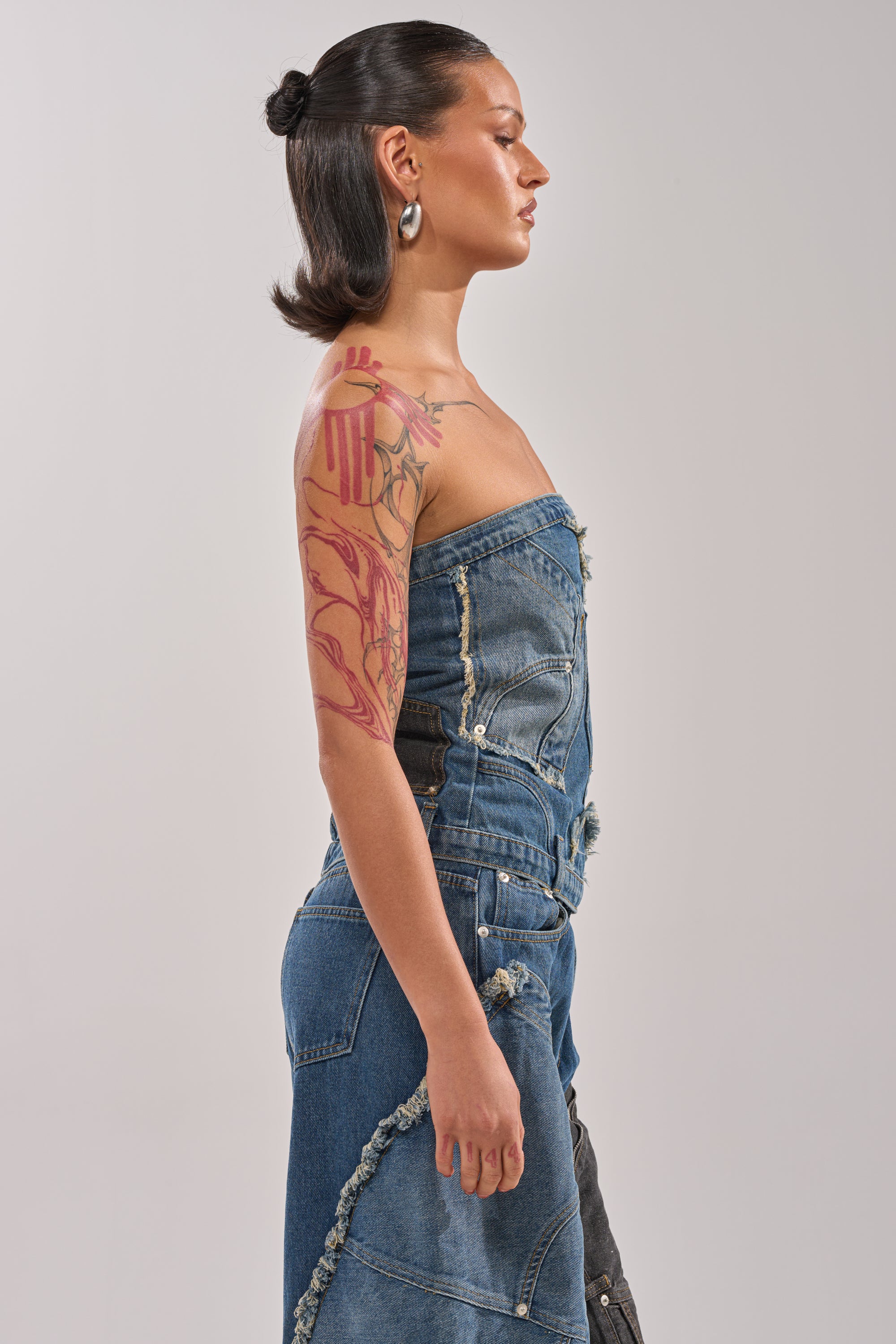 A woman with slicked-back dark hair and hoop earrings stands in profile, embodying street style in the MIXED MATCH DENIM CORSET. The strapless dress with raw edges highlights her colorful tattoo sleeve against a plain light background. 2