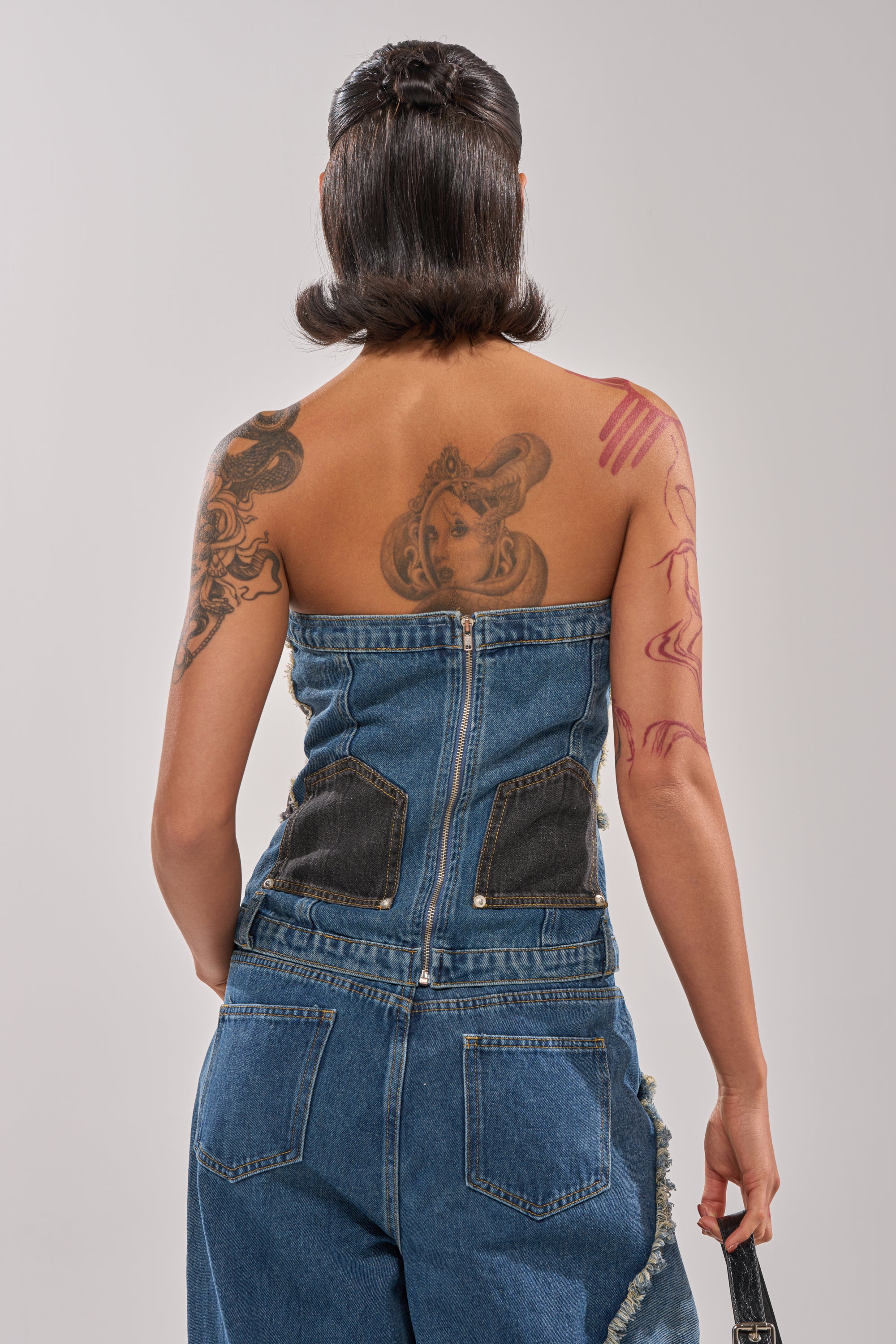 A person with short, dark hair is seen from behind in street style, wearing the MIXED MATCH DENIM CORSET and matching jeans. Their tattooed back and arms feature a large portrait and abstract designs. 3