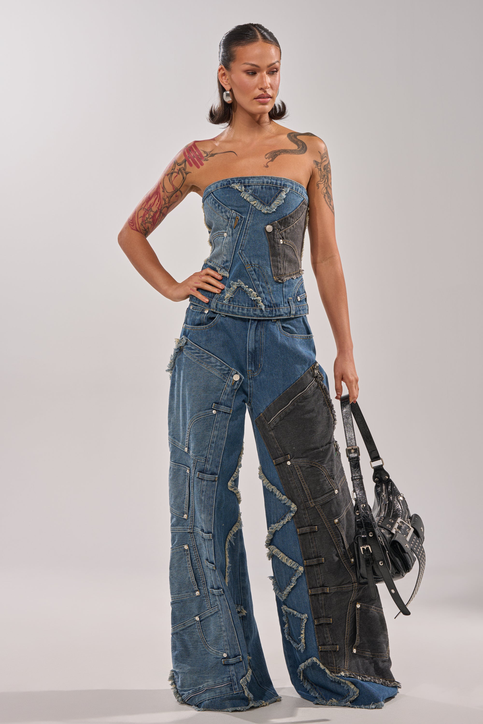 A woman exudes confidence in the MIXED MATCH DENIM CORSET and wide-leg patchwork jeans with frayed edges, showcasing her tattoos and a black handbag against a simple, light background for bold street style. 4