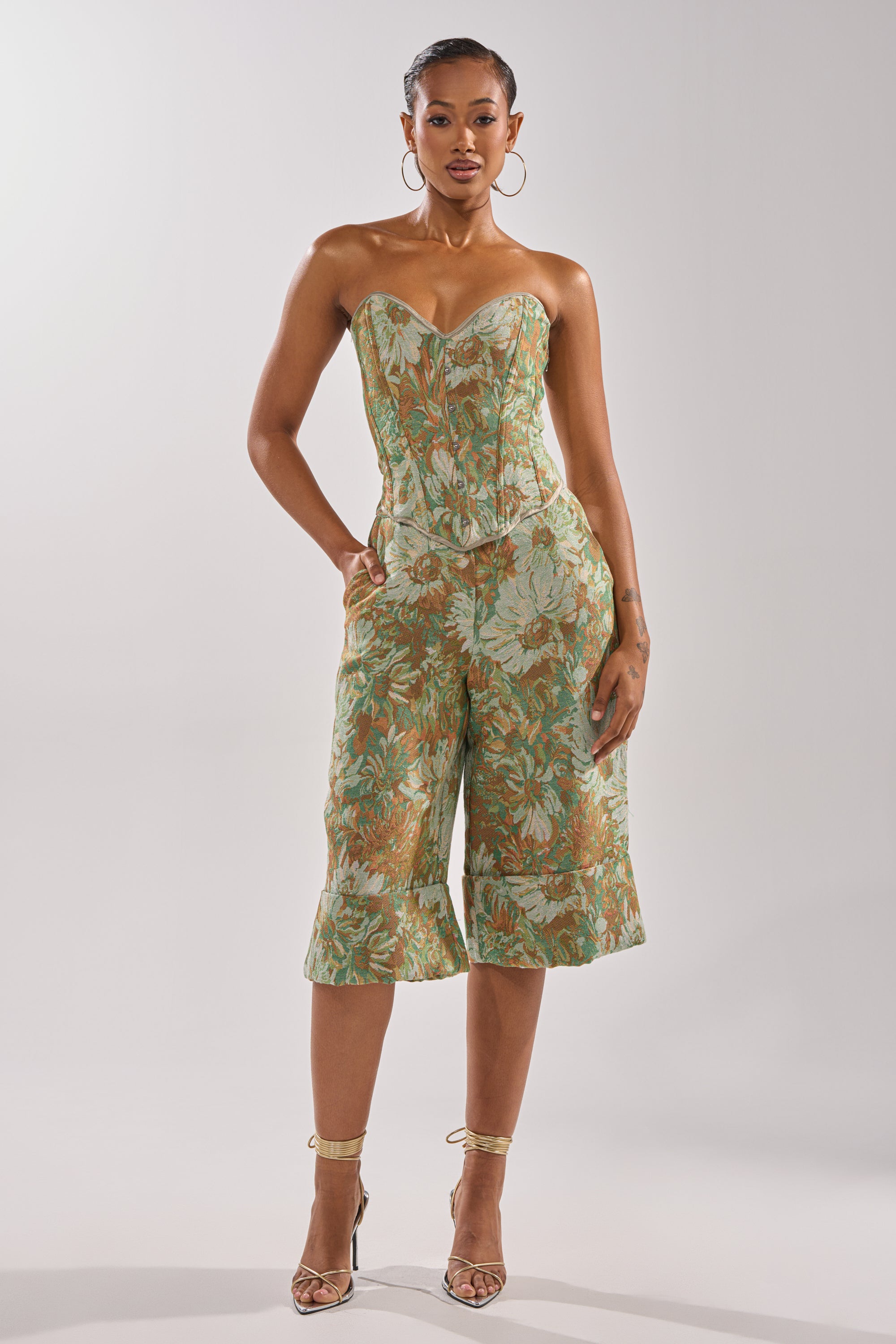 A woman wears the LOVERS FLORAL BERMUDA SHORT in green and orange woven jacquard, paired with a matching strapless floral corset top, hoop earrings, strappy heels, and her hair styled back.