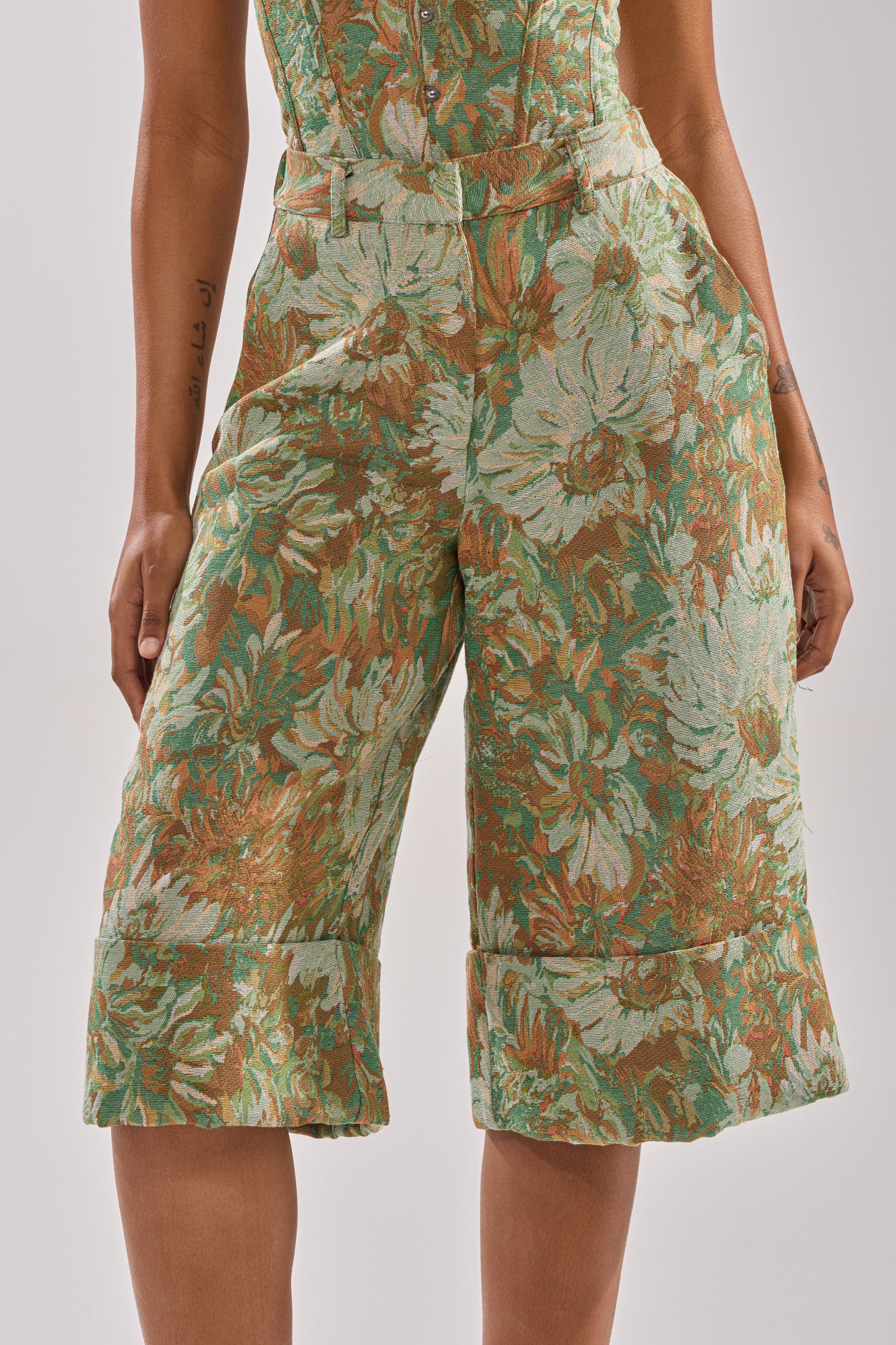 A person wearing the LOVERS FLORAL BERMUDA SHORT, featuring green, brown, and cream floral jacquard fabric, side pockets, and cuffed hems. Only the lower torso and upper legs are visible.