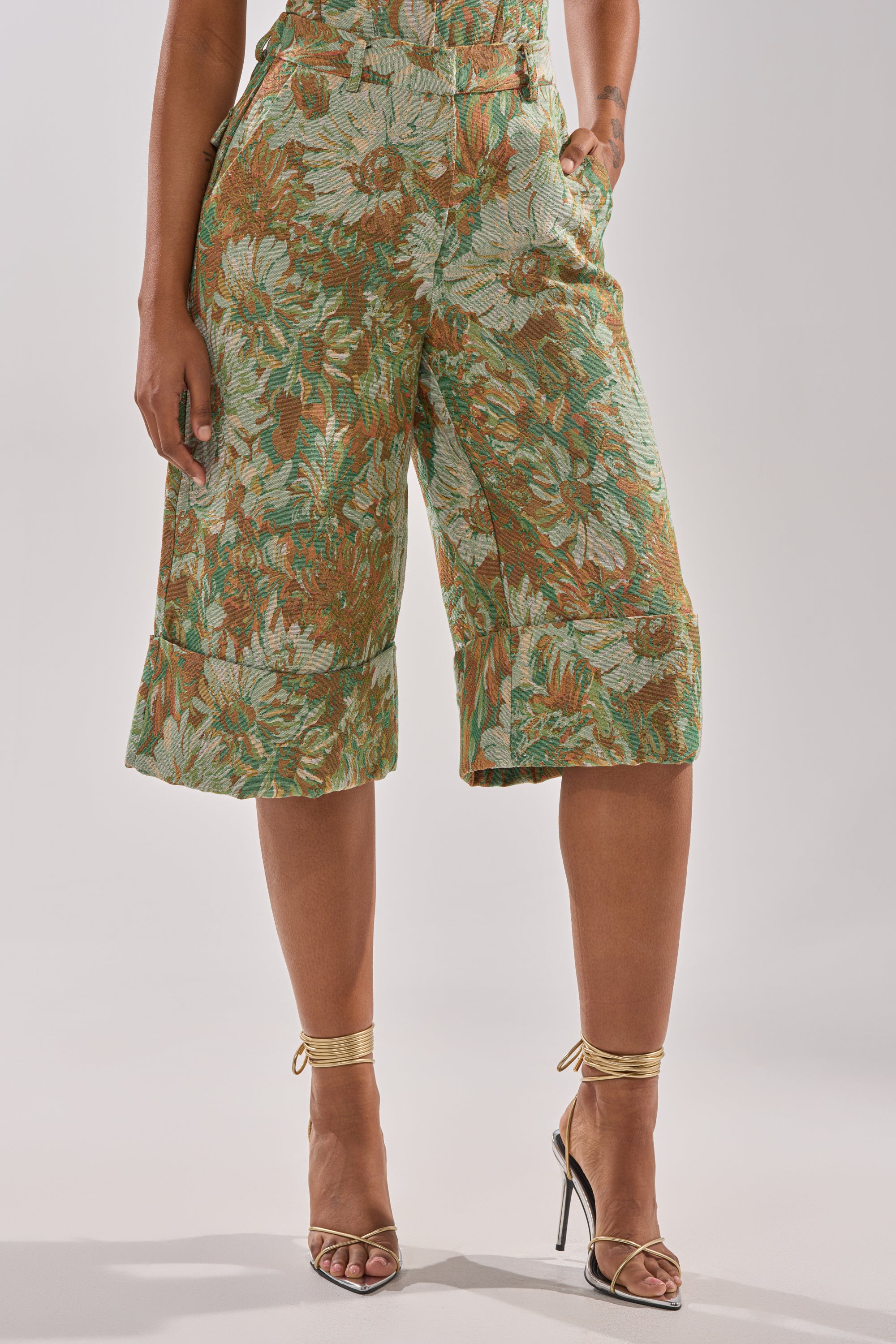 A person wears LOVERS FLORAL BERMUDA SHORTS in green and brown floral jacquard with gold strappy heels, standing against a plain light background. Their hand rests in a pocket, showing only the lower body.