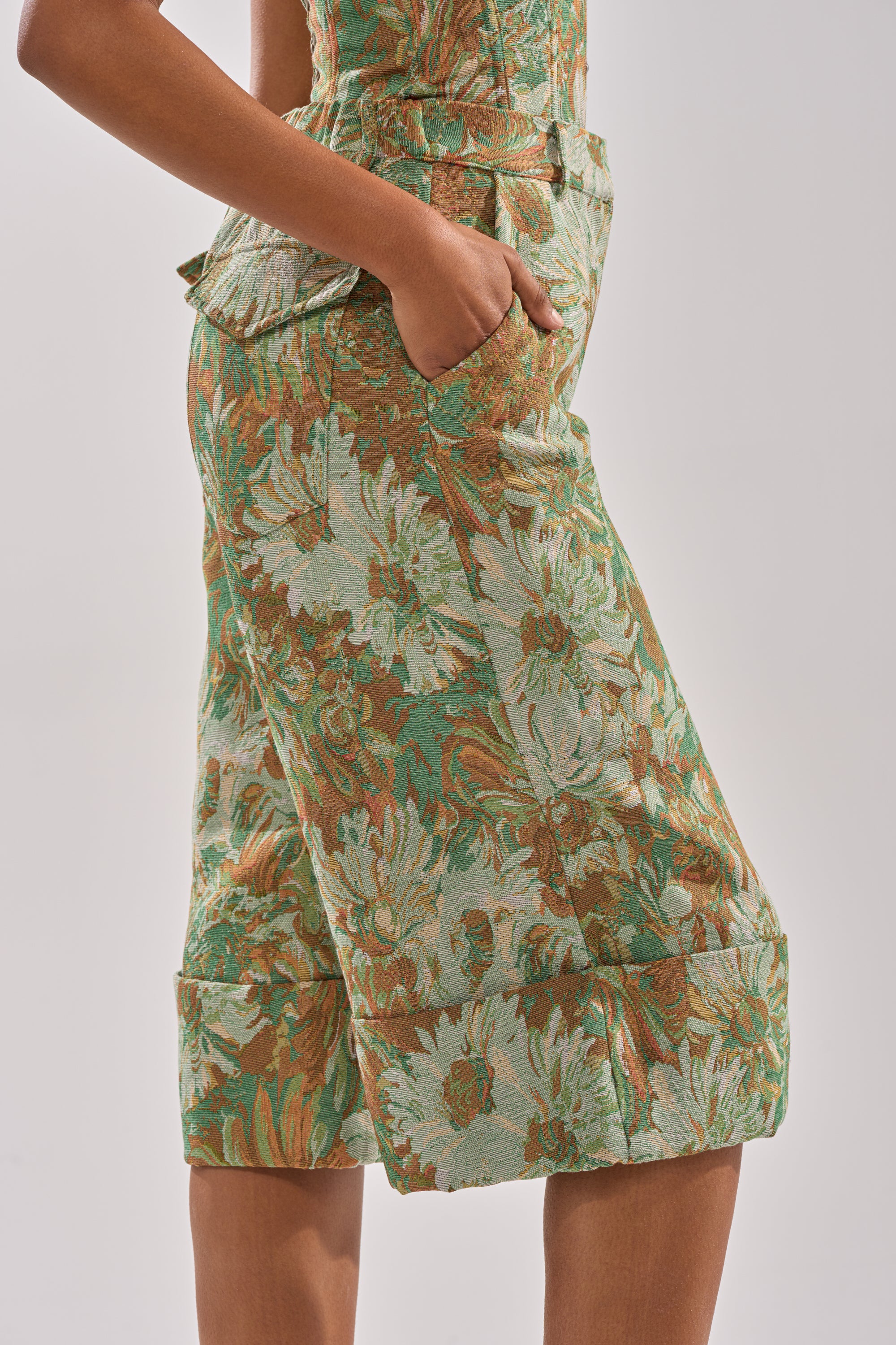 A person models the LOVERS FLORAL BERMUDA SHORT in green and brown jacquard, featuring a floral pattern, posed in profile with one hand in the pocket against a plain background.