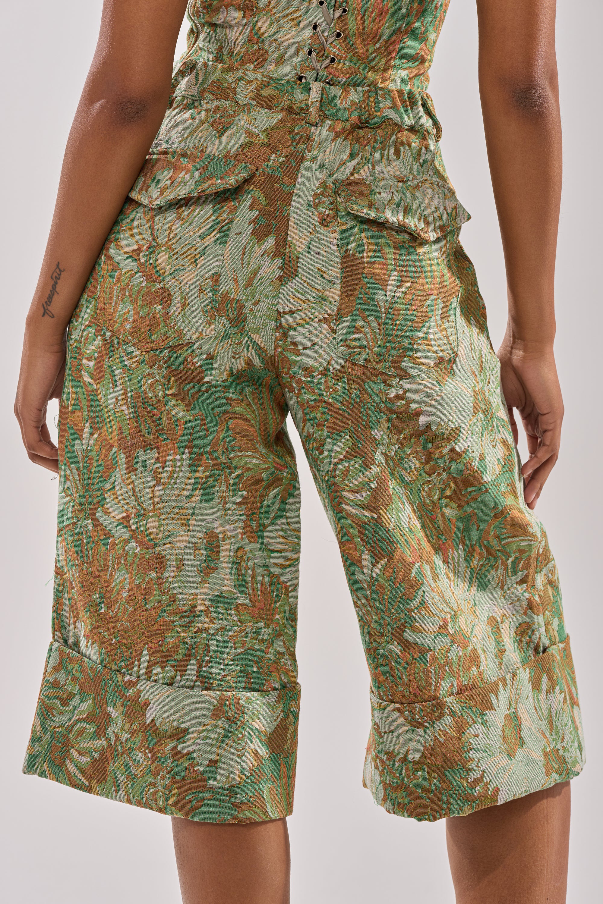 A person shown from the back wears the LOVERS FLORAL BERMUDA SHORT, featuring a green, brown, and beige floral jacquard pattern, high waist, wide-leg fit, flap pockets, and lace-up waist detail.