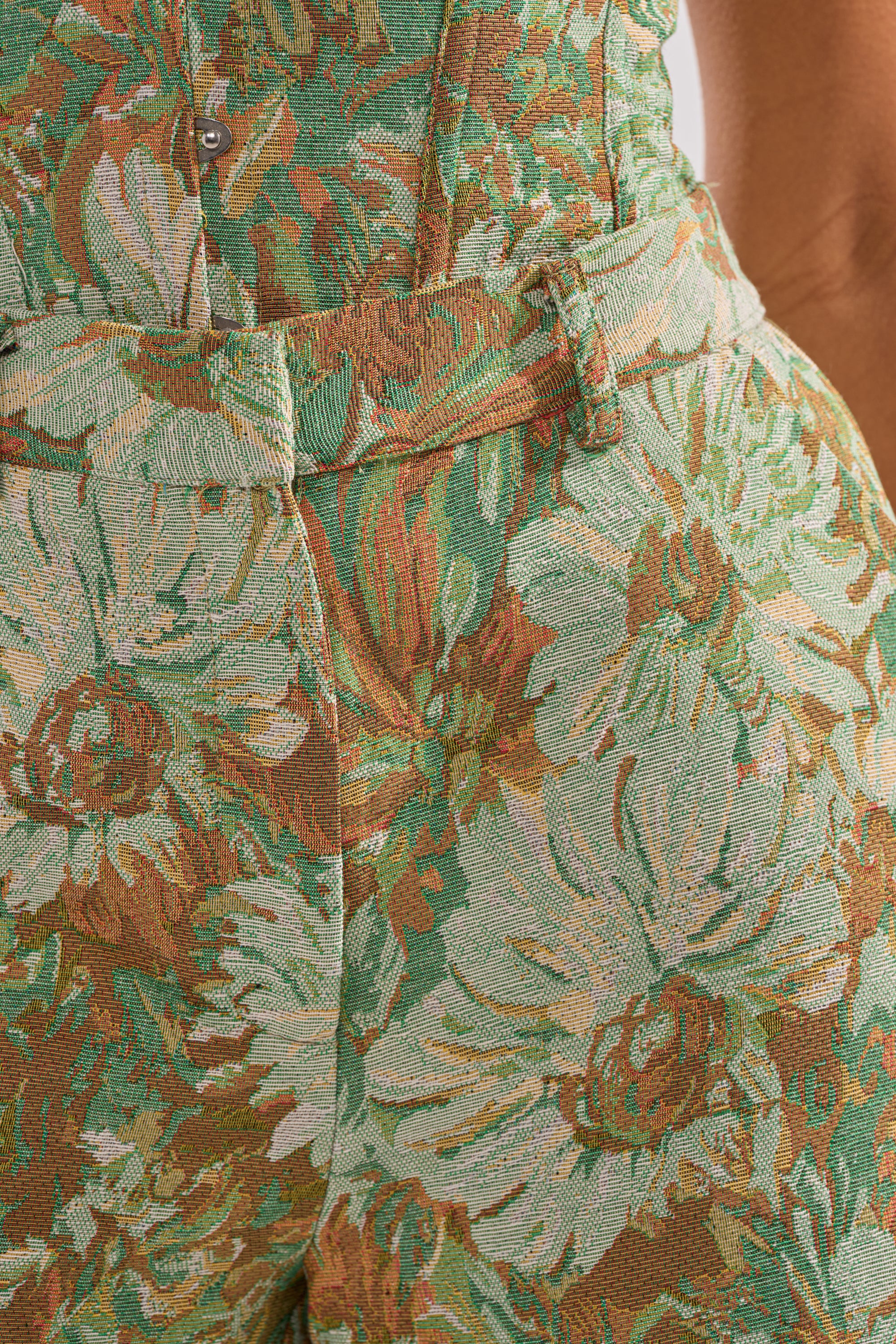 Close-up of a person's waist wearing the LOVERS FLORAL BERMUDA SHORT, crafted from jacquard fabric in green, brown, and cream with large floral and leaf patterns. Features include a waistband with belt loop and visible button.