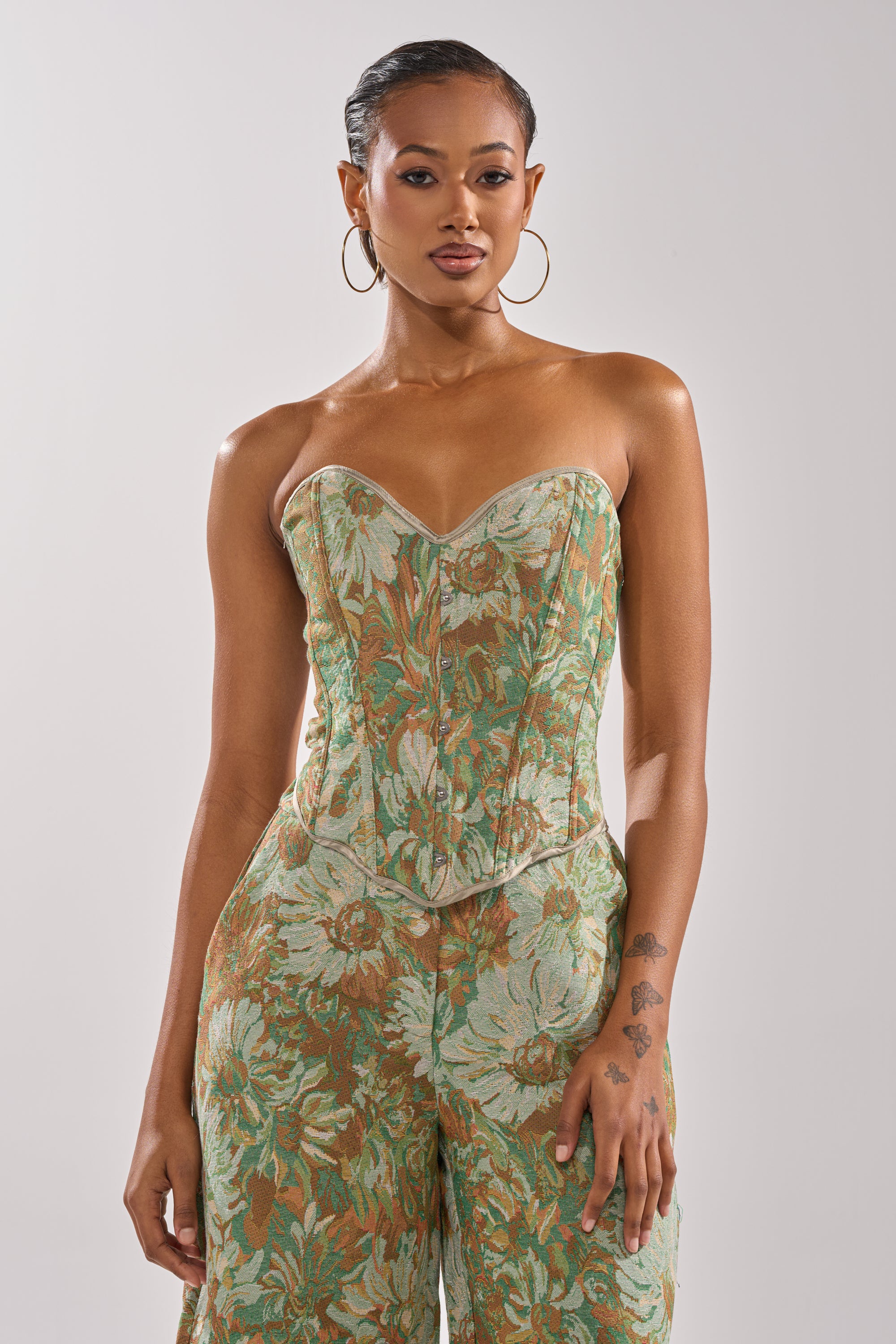 A woman with hoop earrings and a floral forearm tattoo wears the LOVERS FLORAL CORSET jumpsuit featuring a strapless, green floral print with a corset-style bodice and sweetheart neckline, standing confidently against a plain background.