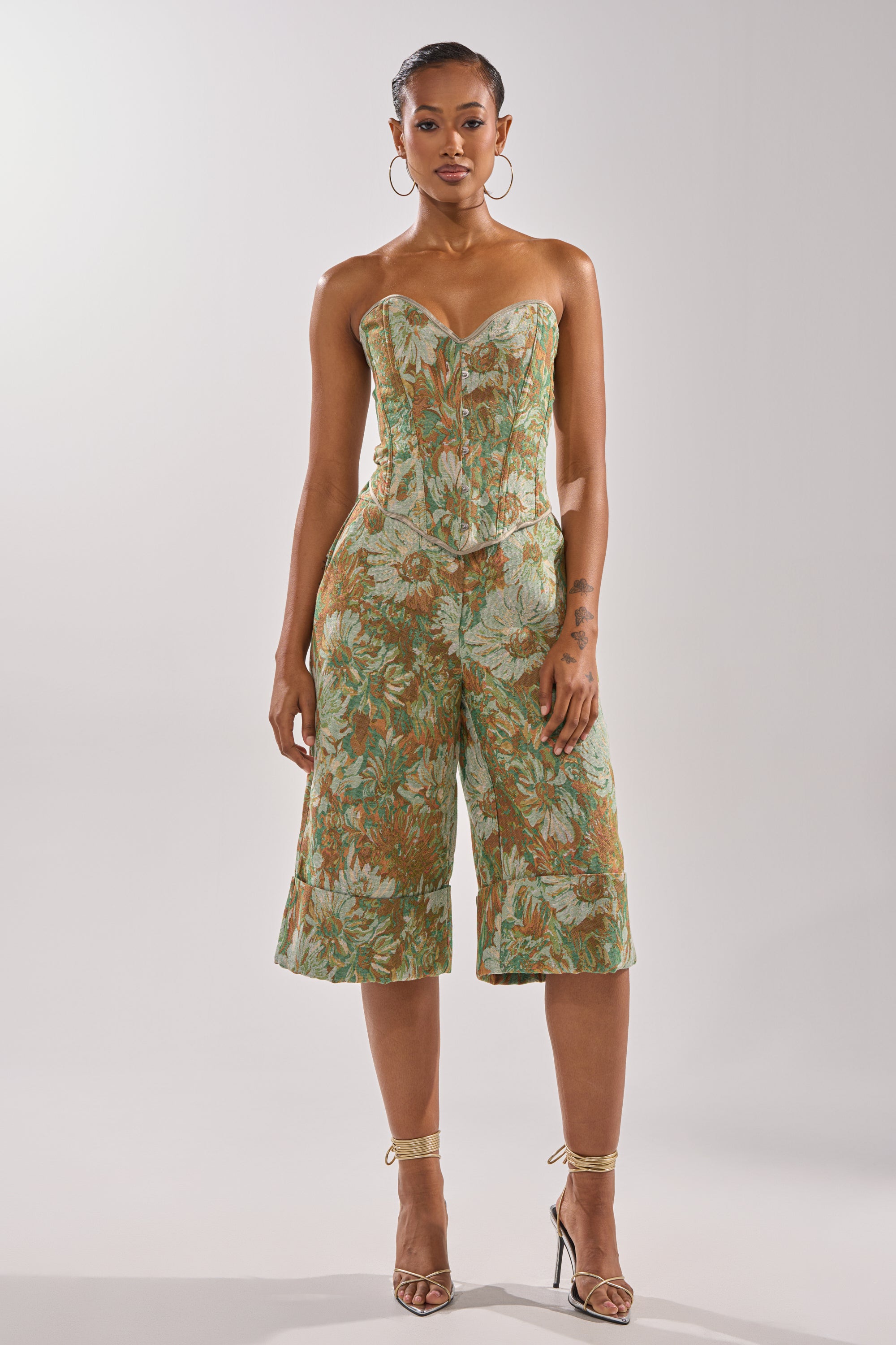 A woman wearing the LOVERS FLORAL CORSET, a strapless green and gold floral top with a sweetheart neckline, paired with matching culotte pants and strappy heels, stands facing forward against a plain light background.