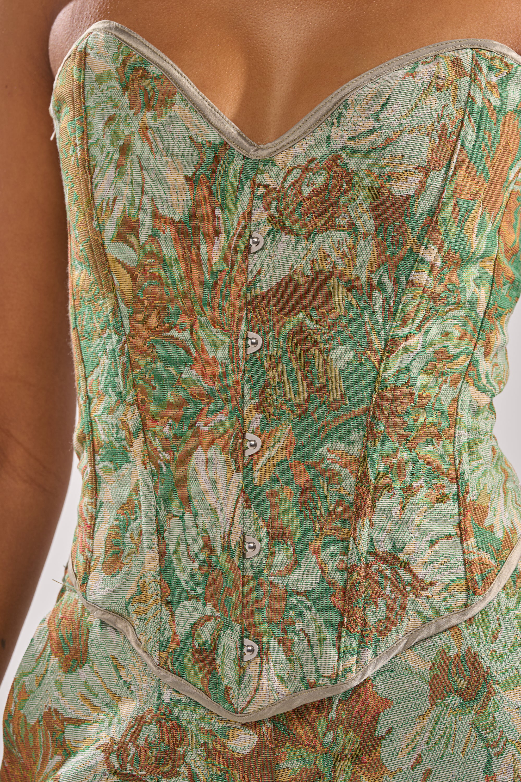 A close-up of a person wearing the LOVERS FLORAL CORSET, featuring a strapless sweetheart neckline, green and brown jacquard floral fabric, silver front buttons, and a textured design for an elegant, vintage-inspired look.