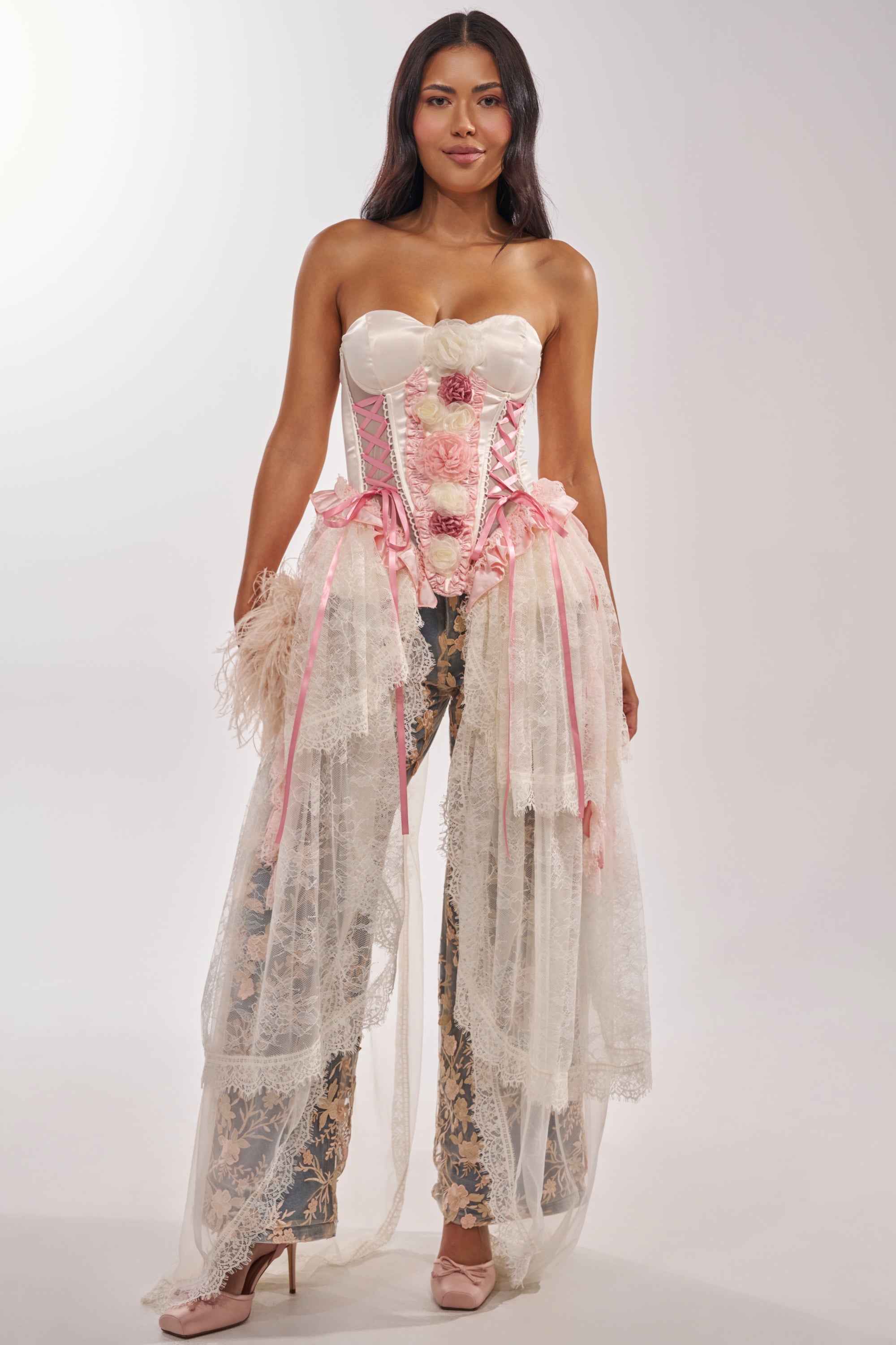 A woman wears the FLY AWAY CORSET, a princess-core white satin top with pink ribbons and floral details, styled with lace-draped trousers and pink heels against a plain light gray background. 1