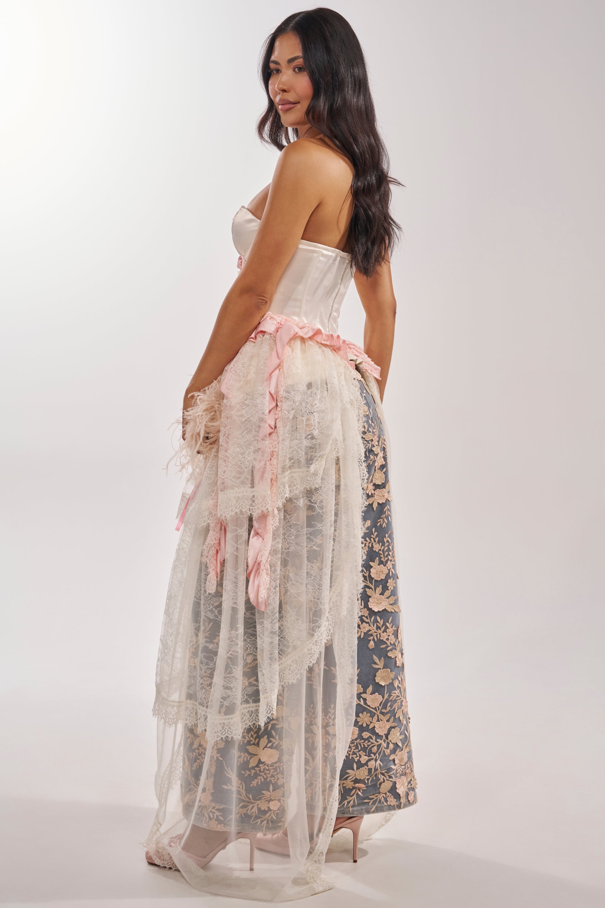 A woman with long dark hair wears the FLY AWAY CORSET dress, featuring a white satin strapless corset top, pink ribbons, lace accents, and a blue floral skirt with sheer white overlay, standing in profile against a plain backdrop. 3
