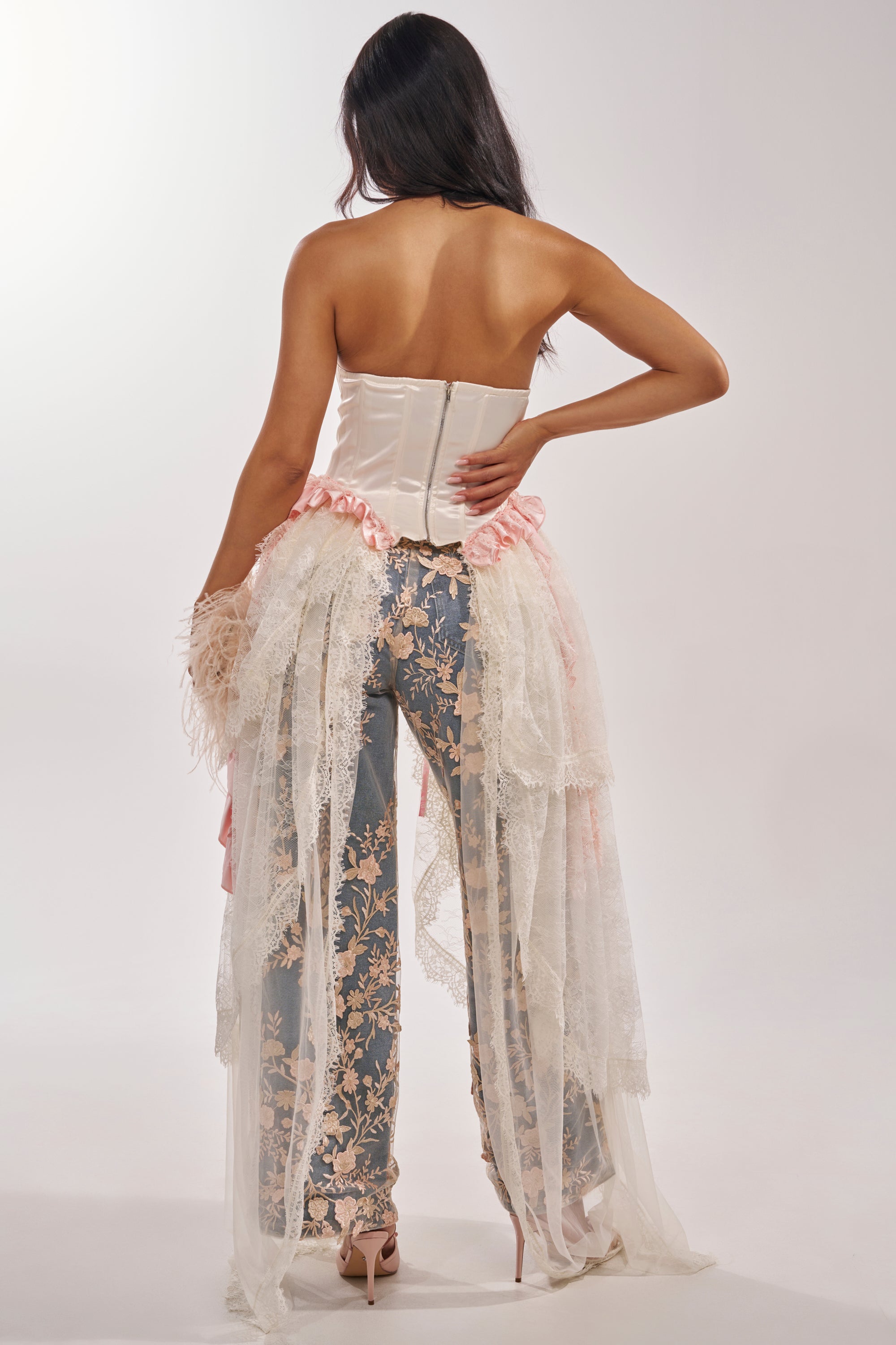 A woman, channeling a princess-core vibe, poses in high heels wearing the FLY AWAY CORSET with blue pants and a sheer floral overlay. Her long dark hair cascades down, complimented by layers of pink and white lace and tulle. 4