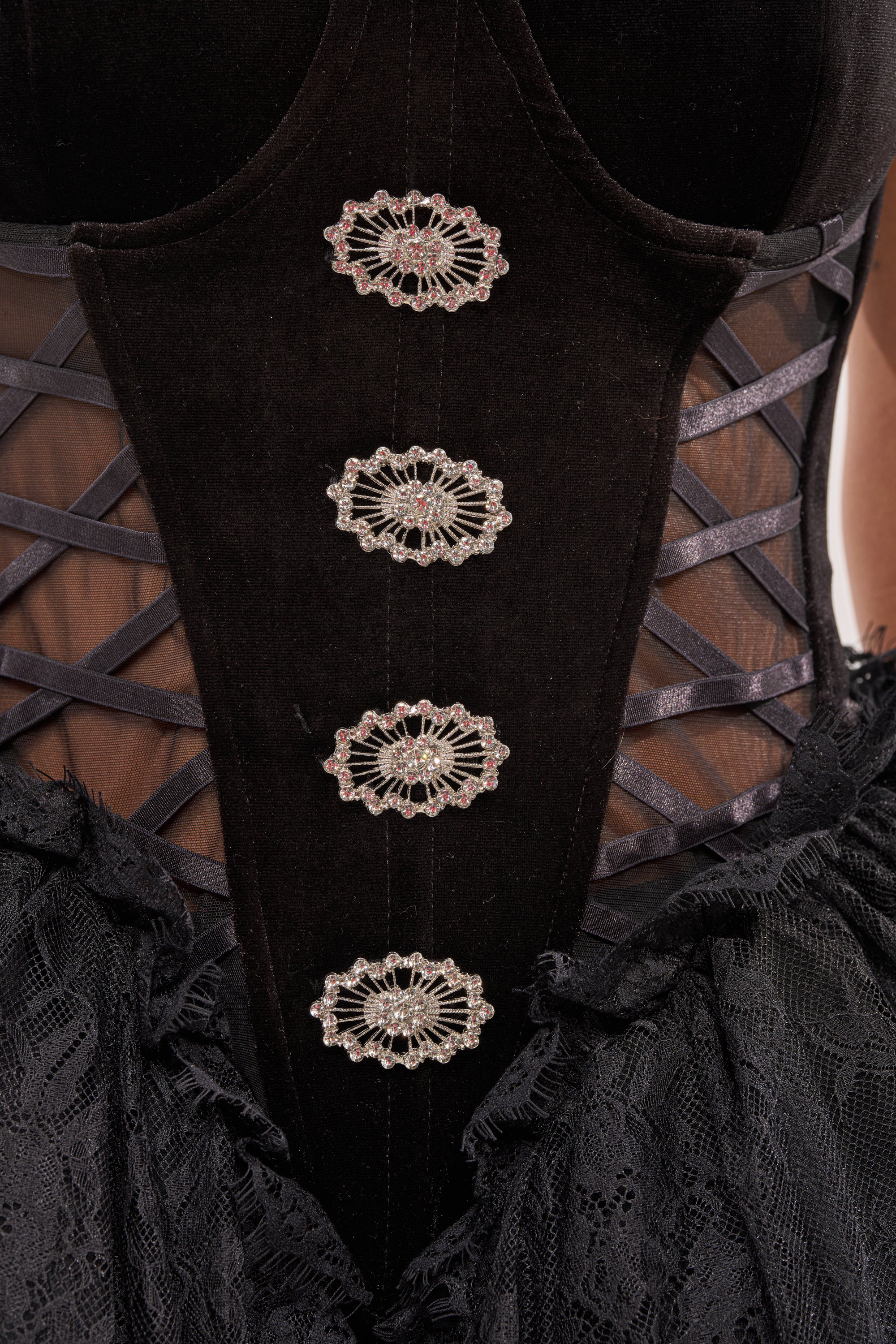 Close-up of the SHE IS DRAMA CORSET in black velvet and lace, showcasing intricate silver brooches, crisscross ribbon details, and rhinestone embellishments on sheer fabric—a dramatic corset for an elegant look. 6
