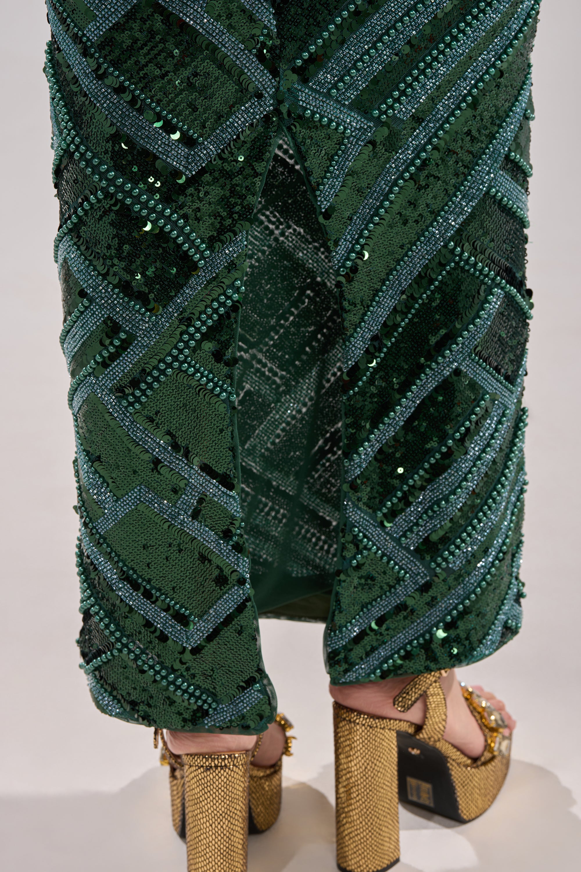 A close-up of a person wearing the HOTTEST IN THE CLUB SEQUIN MAXI SKIRT in emerald green with an intricate geometric pattern, paired with gold textured high-heeled sandals adorned with decorative studs.