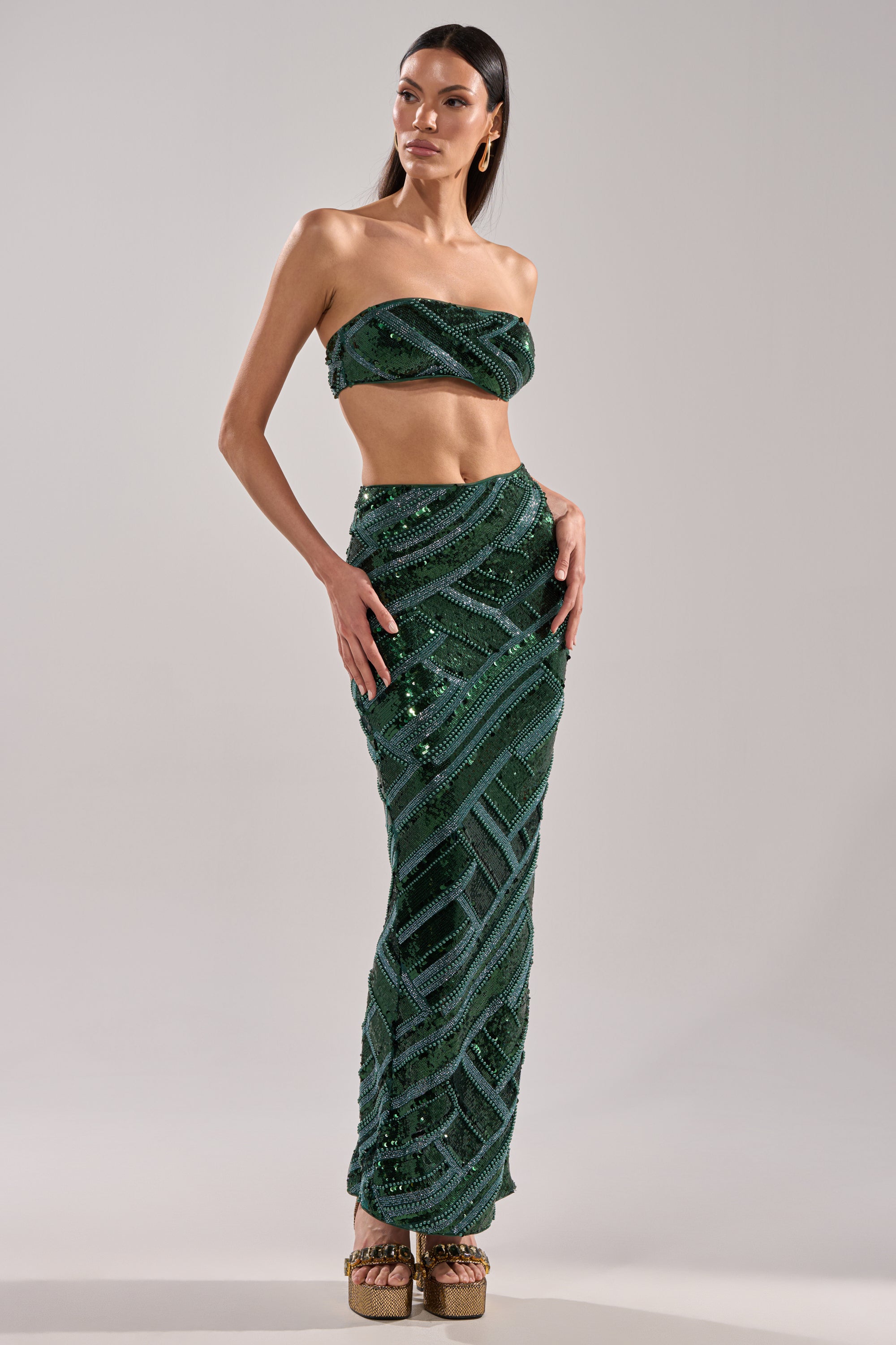 A woman poses in the HOTTEST IN THE CLUB SEQUIN MAXI SKIRT, a high-rise fit maxi skirt with dark green sequins and geometric patterns, paired with a strapless sequin crop top and platform heels against a light gray background.