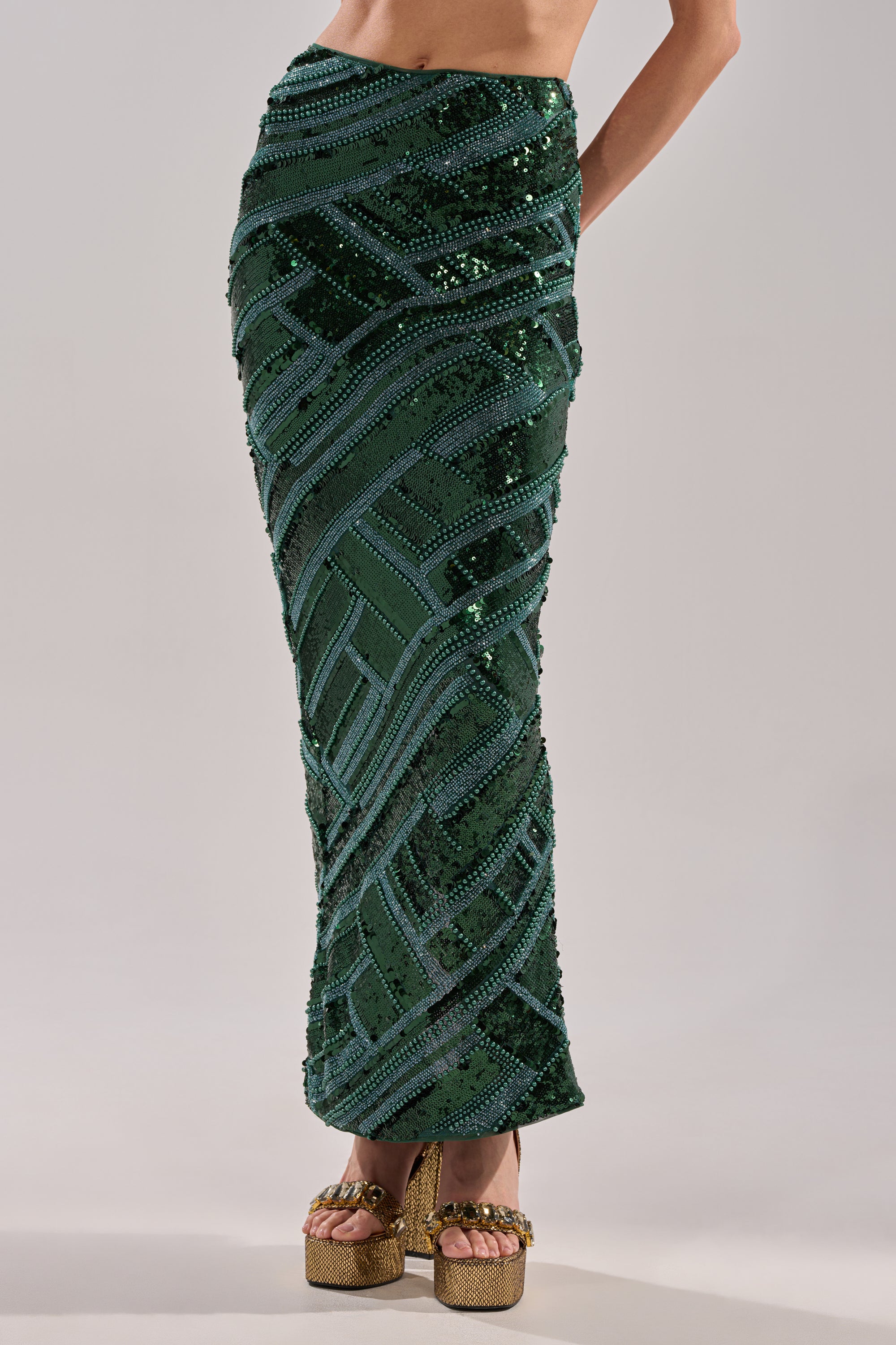 Wearing the HOTTEST IN THE CLUB SEQUIN MAXI SKIRT—a high-waisted, floor-length, dark green geometric sequin skirt—paired with gold platform heels. The bold clubwear look highlights the skirt; upper torso and face are not shown.