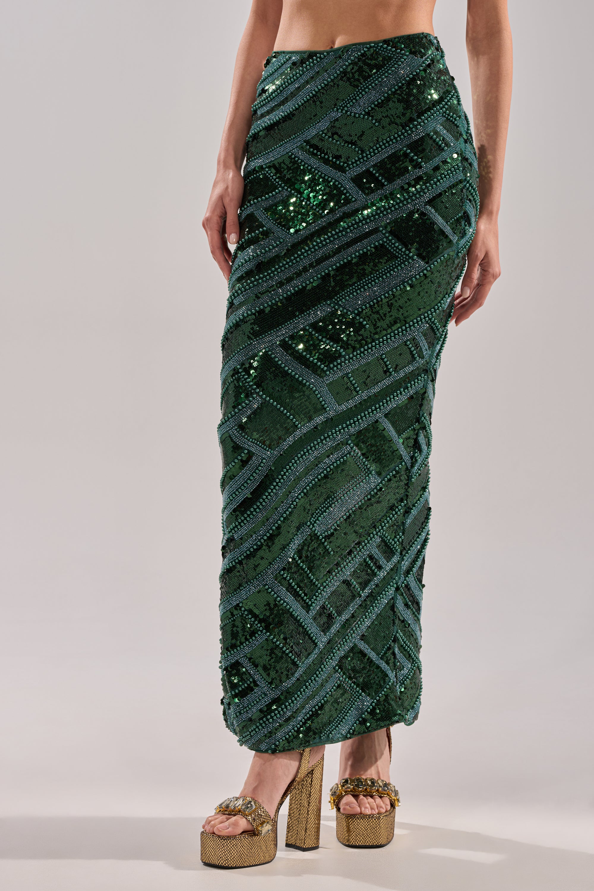 Someone stands against a plain background wearing the HOTTEST IN THE CLUB SEQUIN MAXI SKIRT, featuring dark green sequins in a geometric pattern and a mermaid silhouette, paired with gold high-heeled platform sandals.