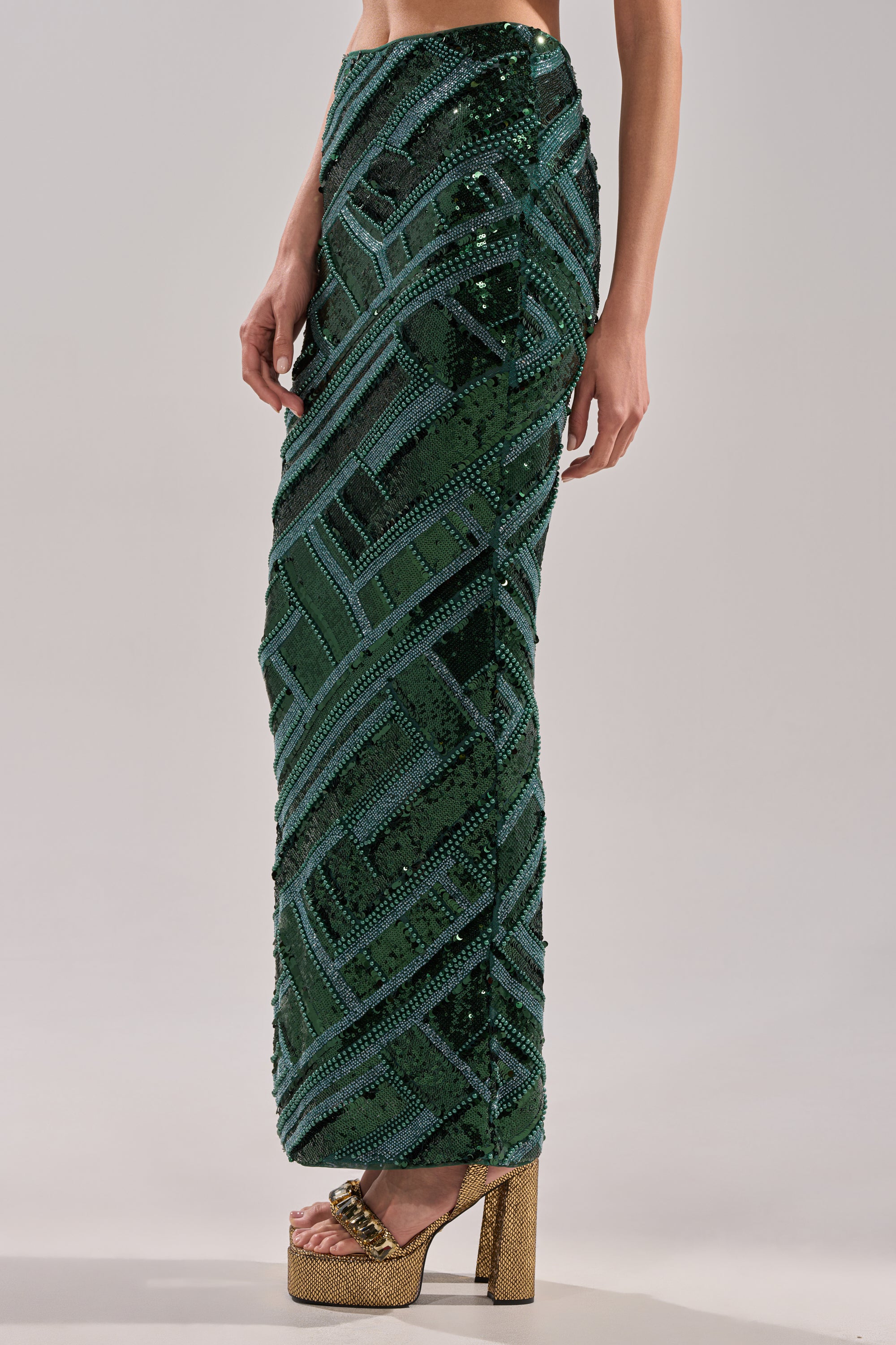 A model wears the HOTTEST IN THE CLUB SEQUIN MAXI SKIRT, a green high-waisted maxi with geometric sequins and a floor-length mermaid silhouette, paired with gold platform heels against a plain light grey background.